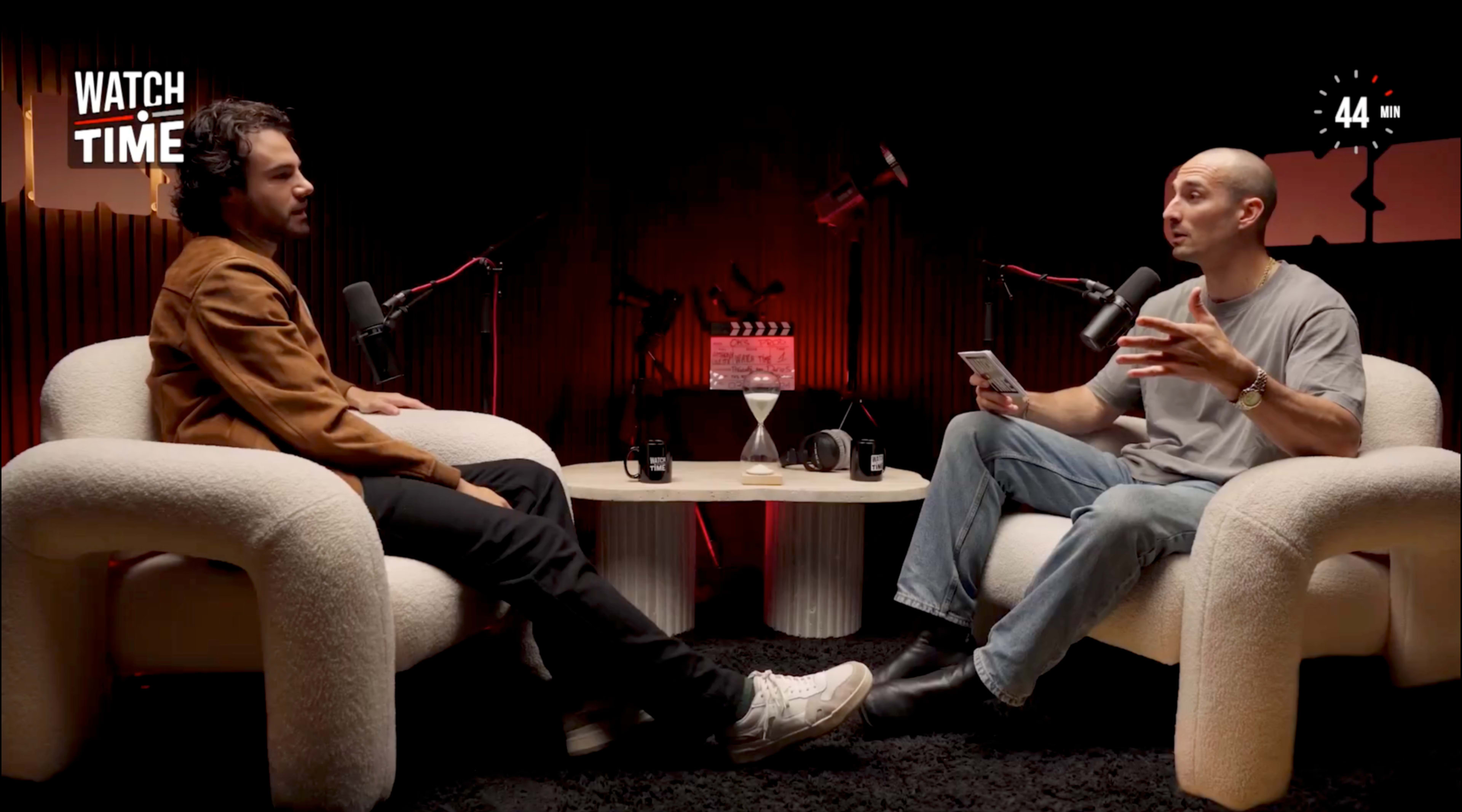 Two individuals are seated in modern armchairs in a dimly lit studio, engaged in conversation while microphones are positioned in front of them.