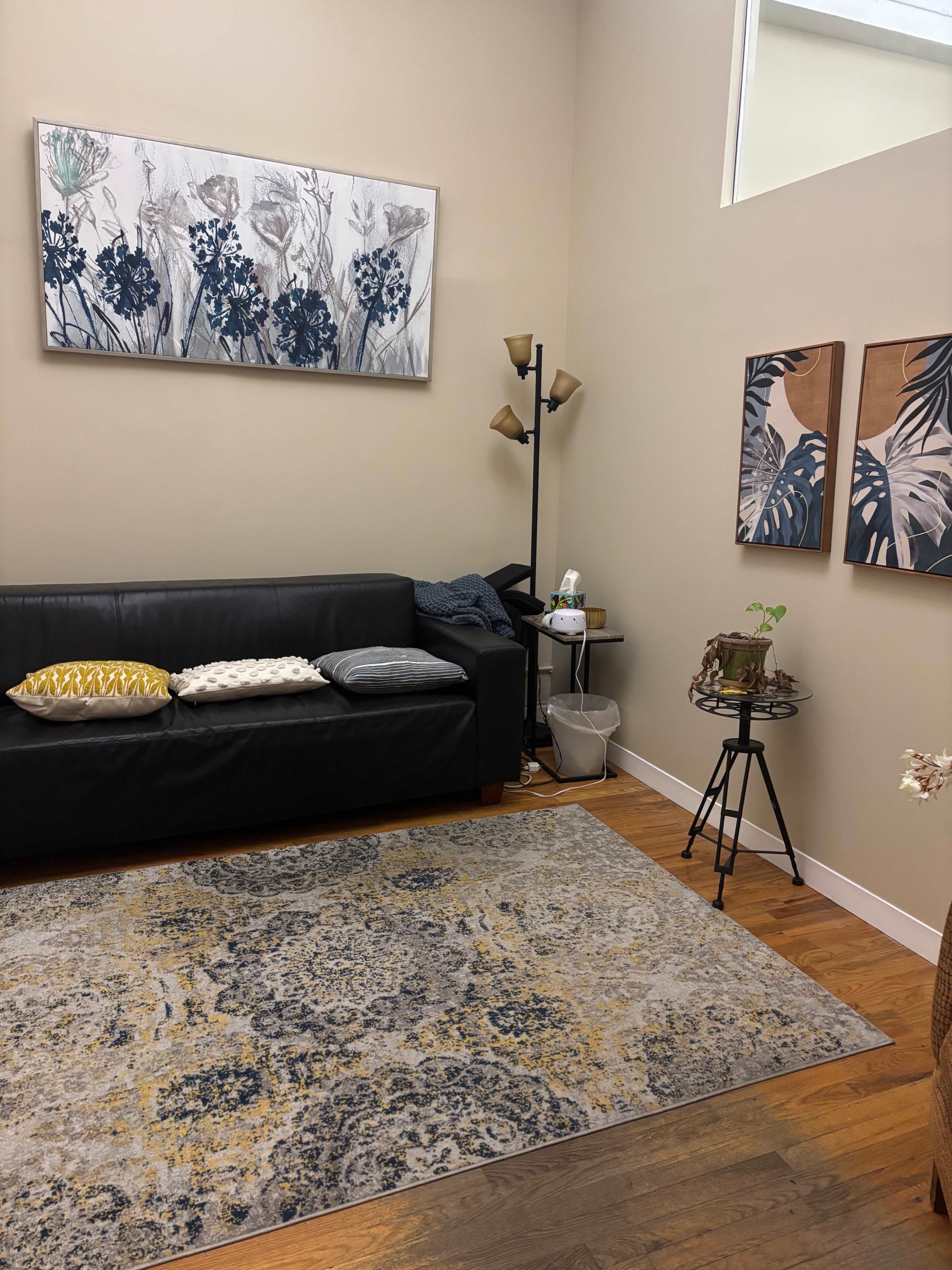 The image shows a cozy living room with a black sofa, decorative pillows, two wall paintings, a standing lamp, and a small plant on a metal side table.