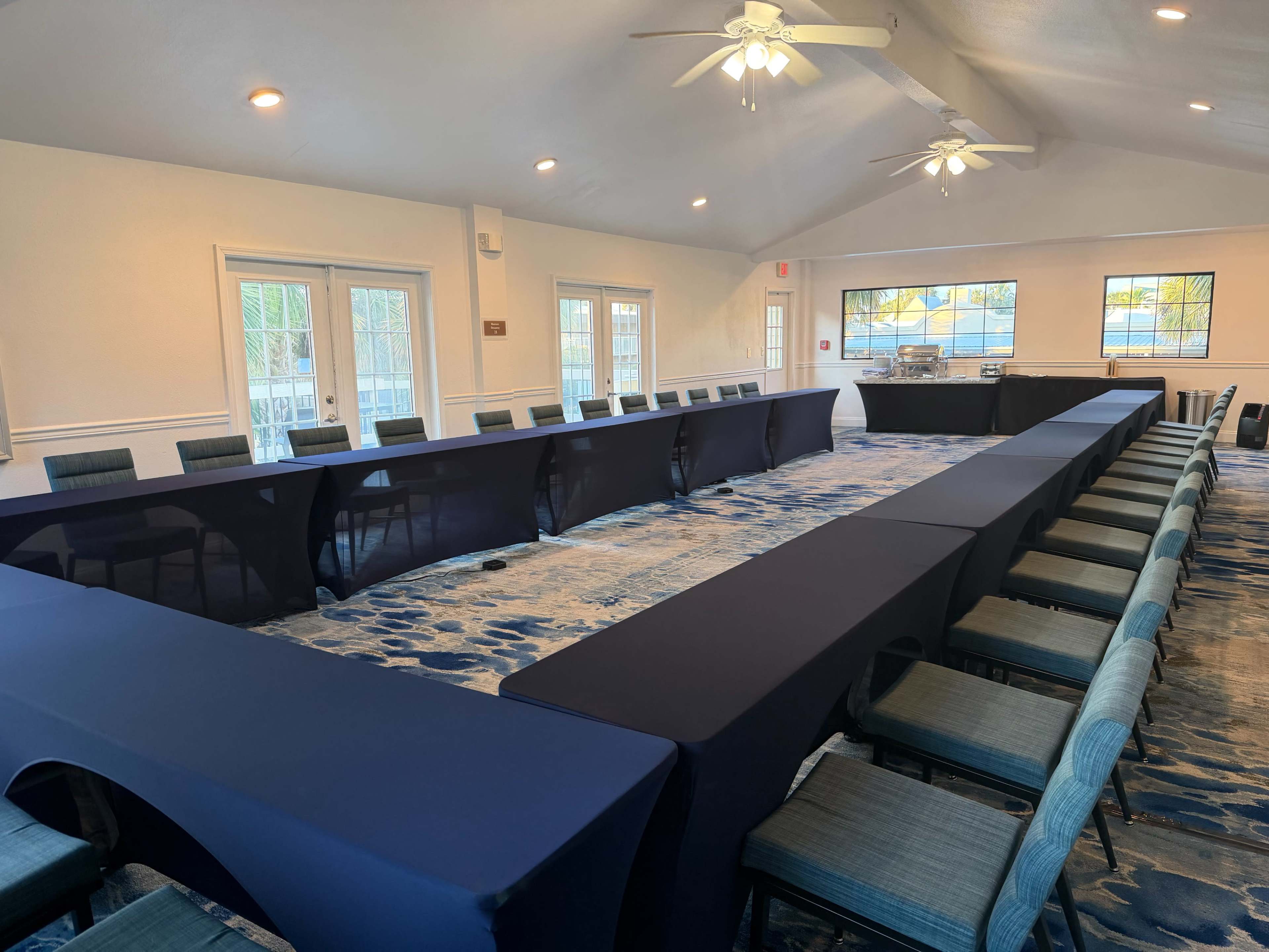 The image shows a conference room arranged with multiple tables covered in blue tablecloths, forming a large U-shape, with chairs positioned around them.