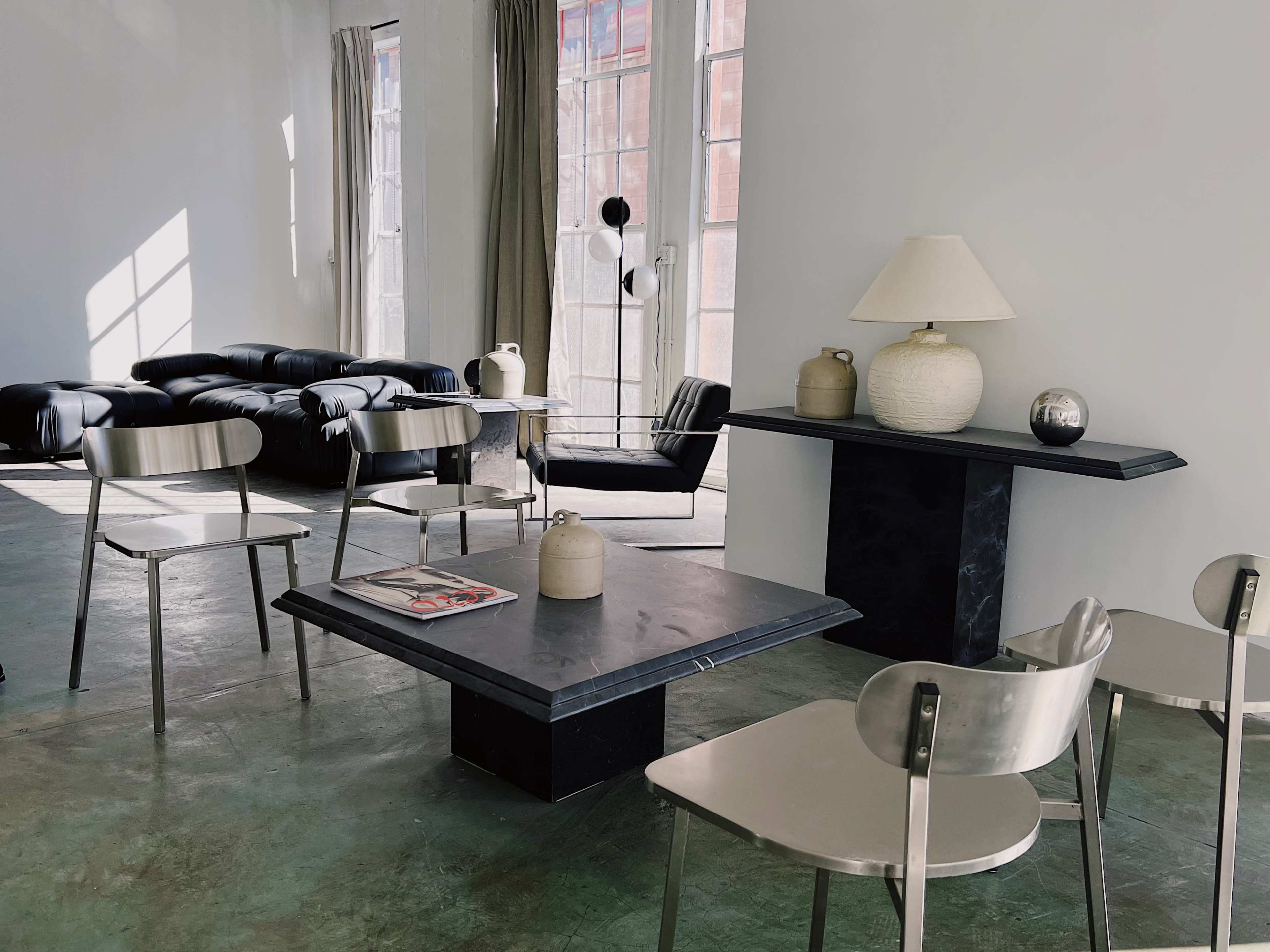 A modern, minimalist living room featuring a black leather sofa, a square coffee table, metal chairs, and decorative objects, all set against a light-colored wall with large windows.