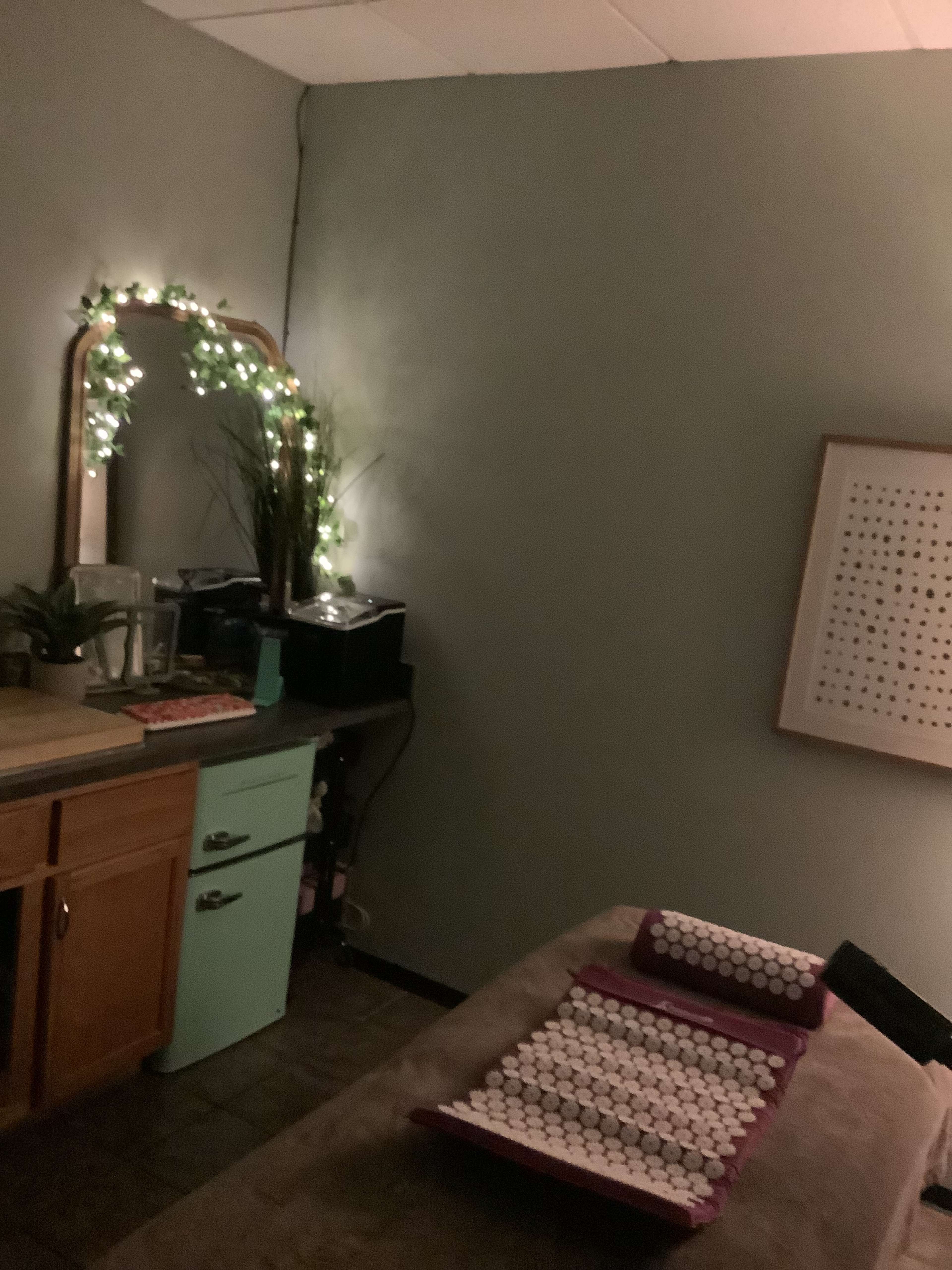 The image shows a dimly lit therapy room with a treatment table, a mirror adorned with lights, and a green cabinet against the wall.