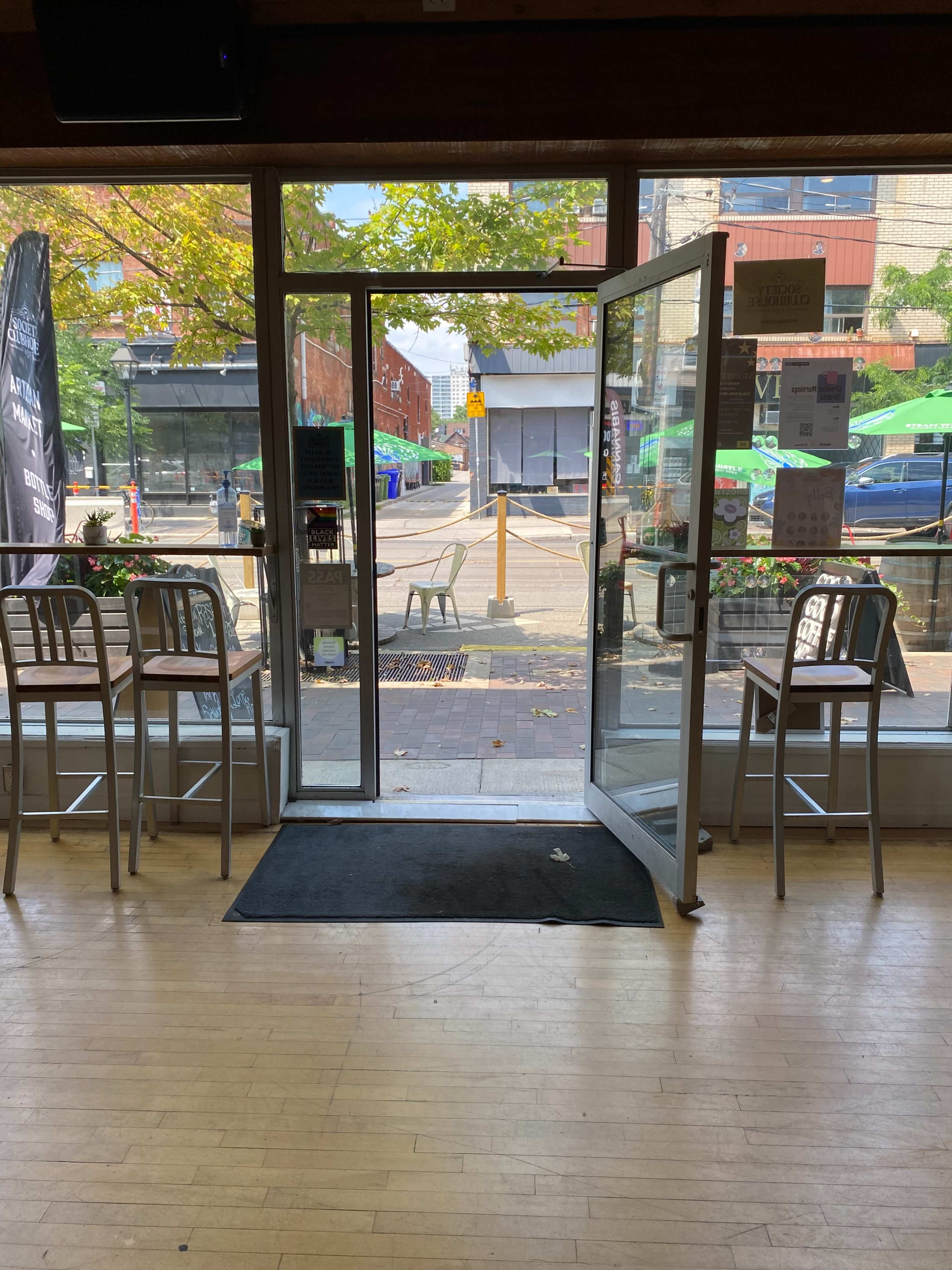 An open glass door leading to a street, with empty bar stools positioned around a small table inside the establishment.