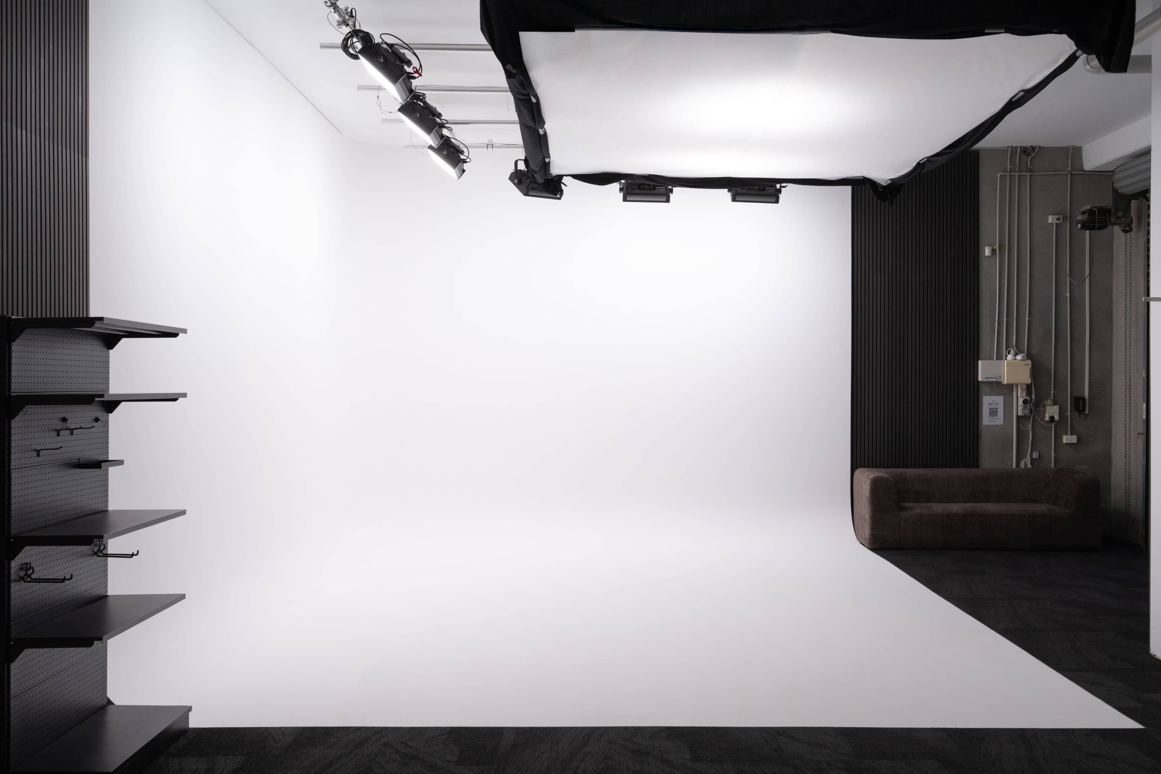 The image shows a minimalist photo studio with a white backdrop, lighting fixtures, and a brown couch in the corner.