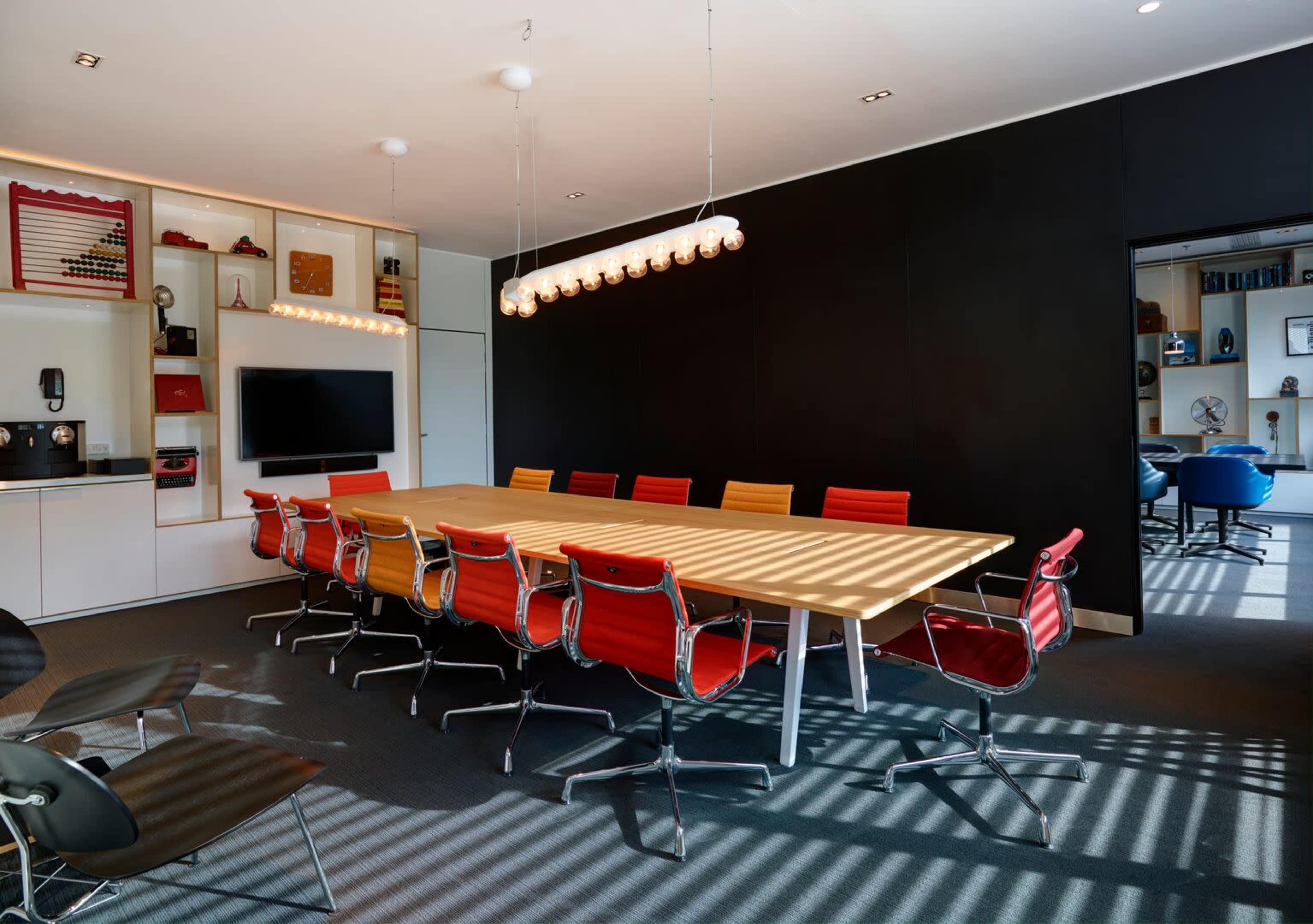 A modern conference room features a long wooden table surrounded by colorful chairs and is illuminated by overhead pendant lights.