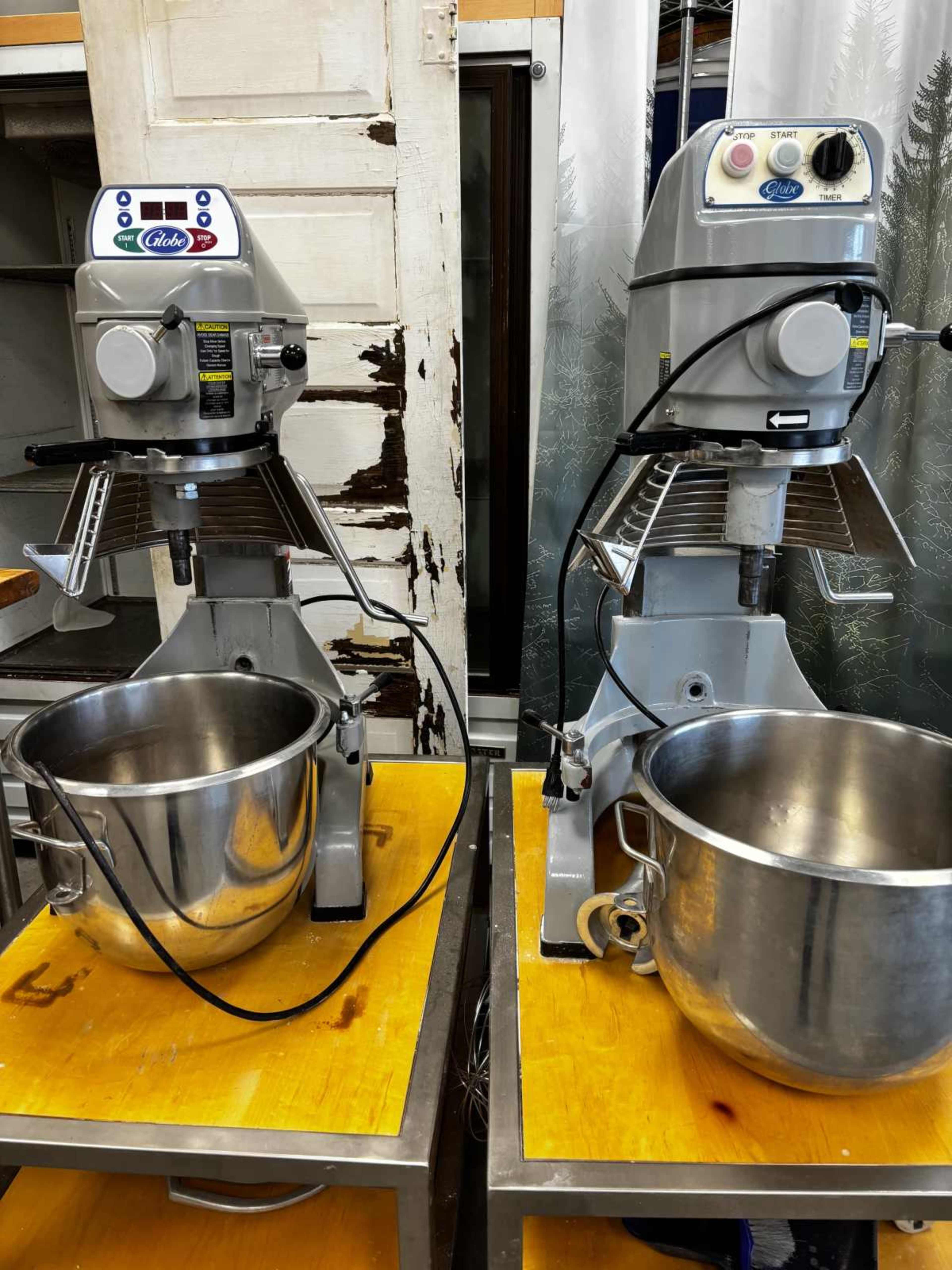 The image shows two commercial stand mixers on wooden tables, each with a large stainless steel mixing bowl and various controls on the front.