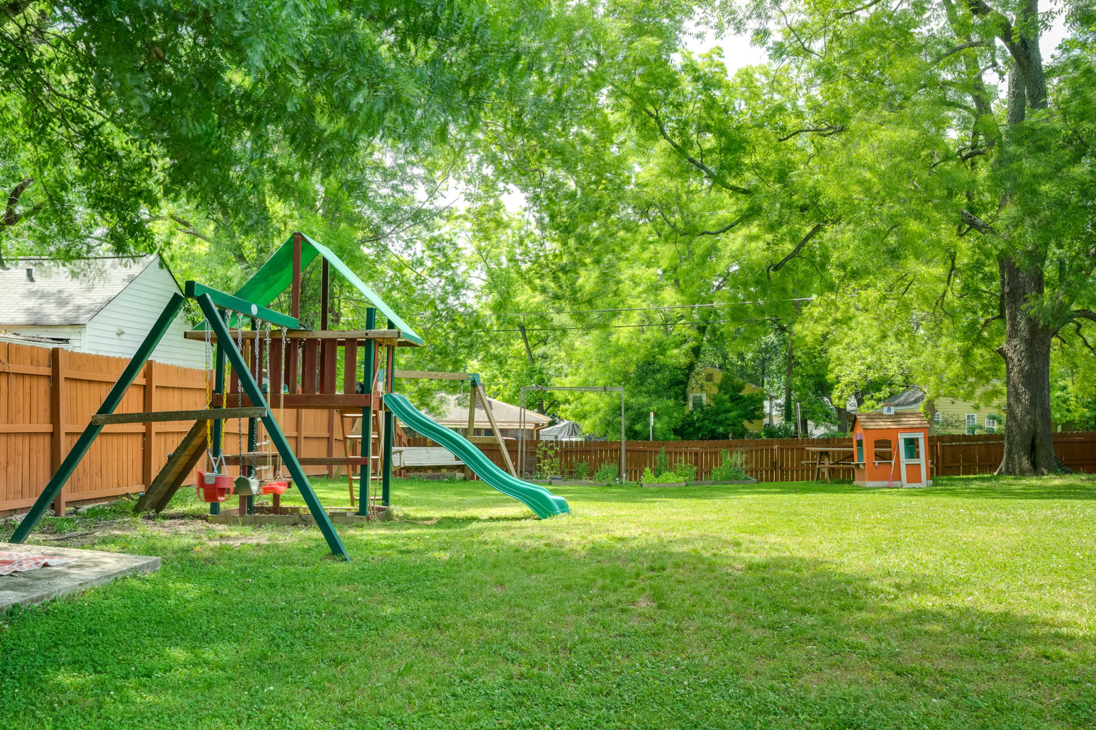 A backyard with a playground structure that includes a slide and swings, next to a small playhouse, all surrounded by green grass and tall trees.