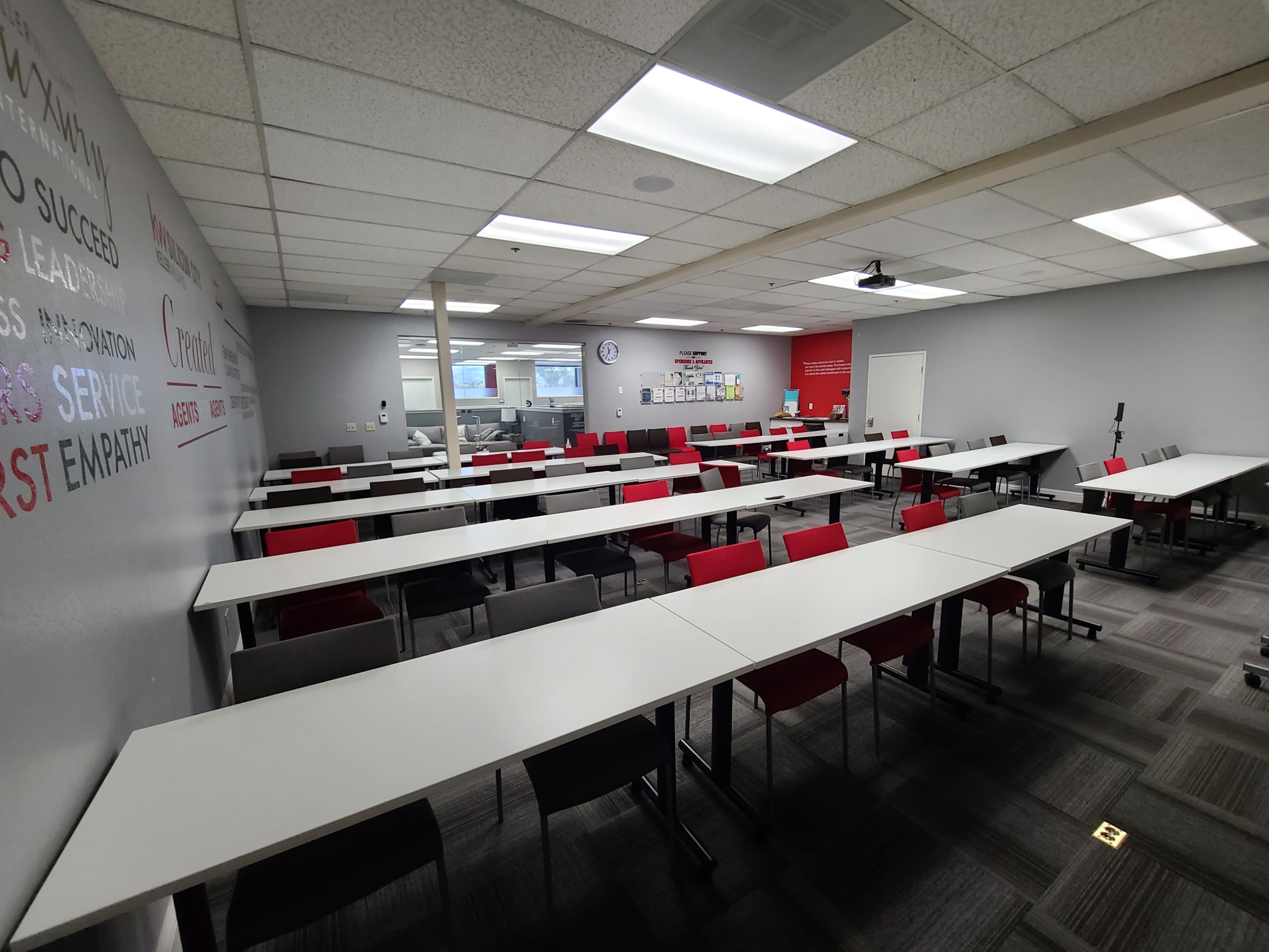 A classroom with multiple tables arranged in rows, equipped with chairs, a whiteboard, and a clock on the wall.