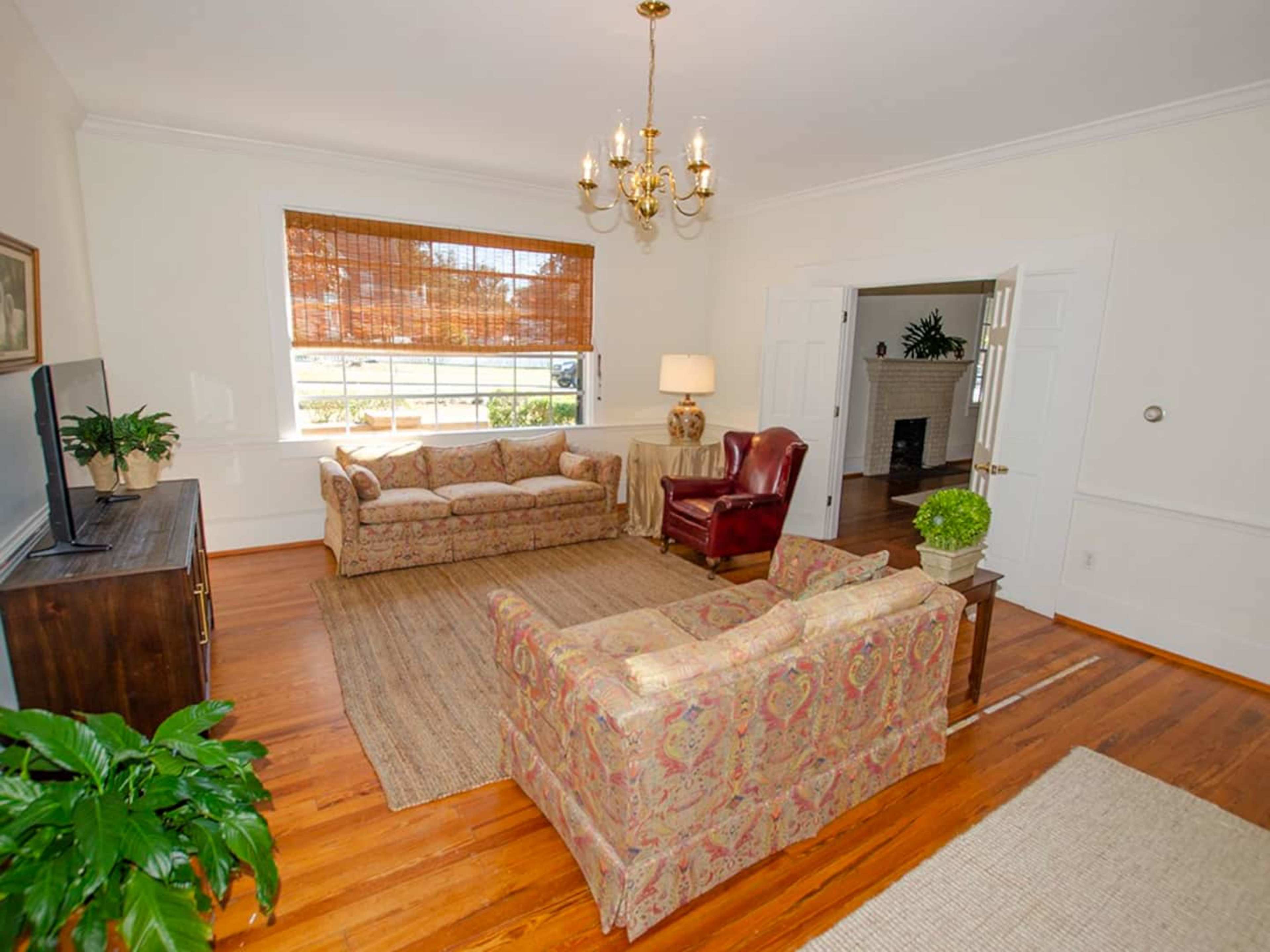 The living room features two sofas, a leather armchair, a television console, and large windows with bamboo shades, set on hardwood floors.