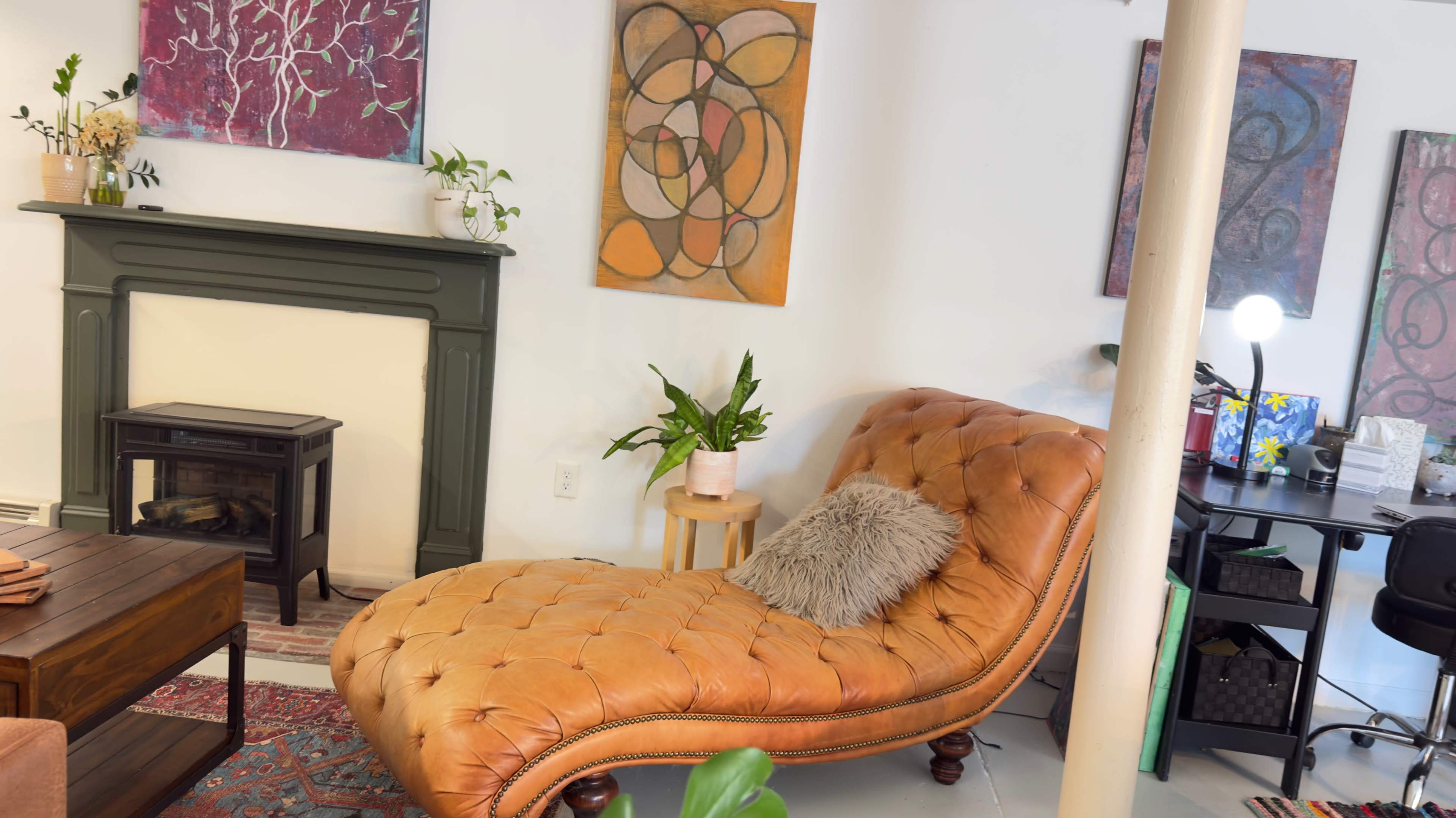 The image depicts a cozy living space featuring a tufted orange chaise lounge, a small indoor plant beside it, and colorful abstract artwork on the walls.