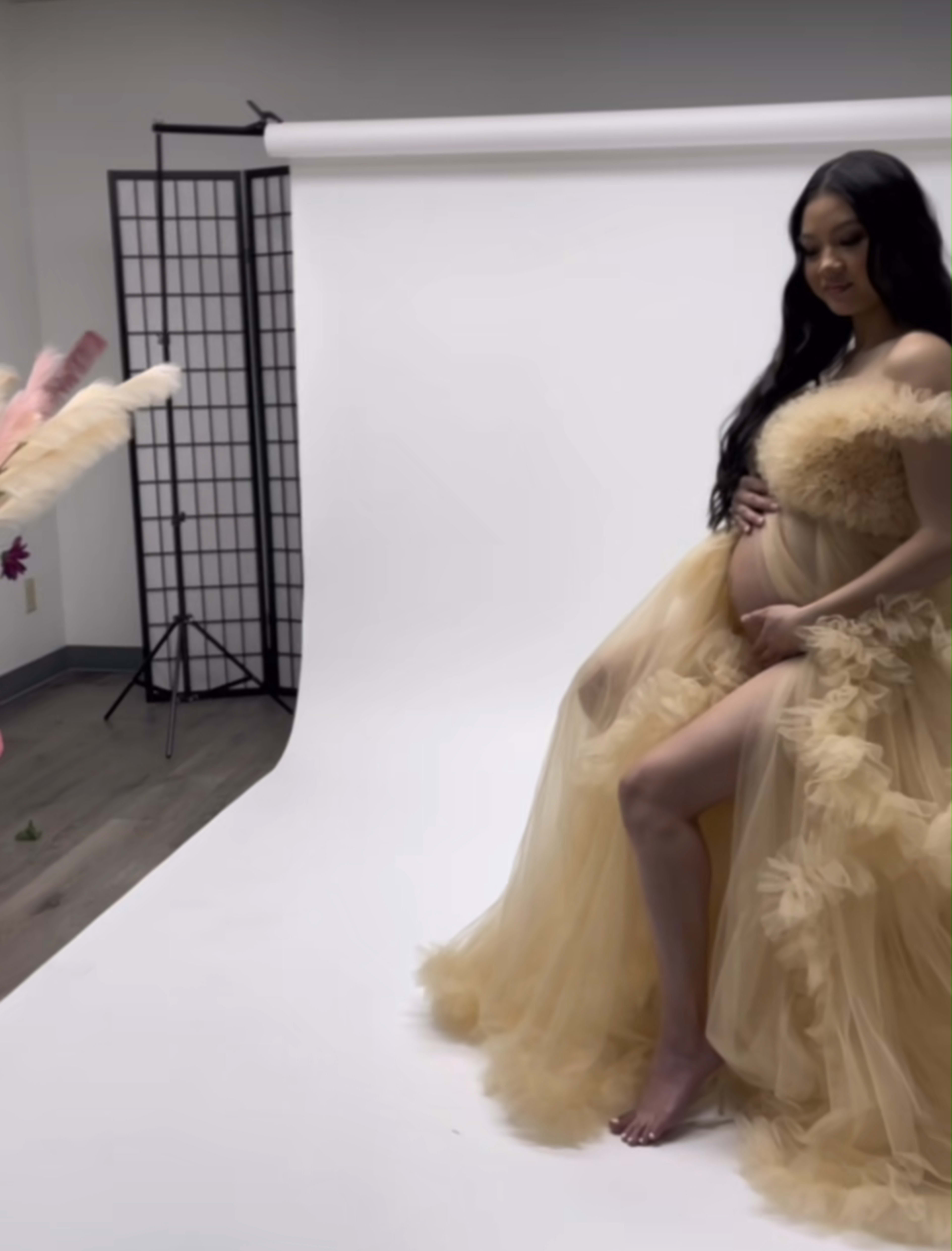 A pregnant model poses in a flowing, nude-colored gown against a plain backdrop in a studio setting.
