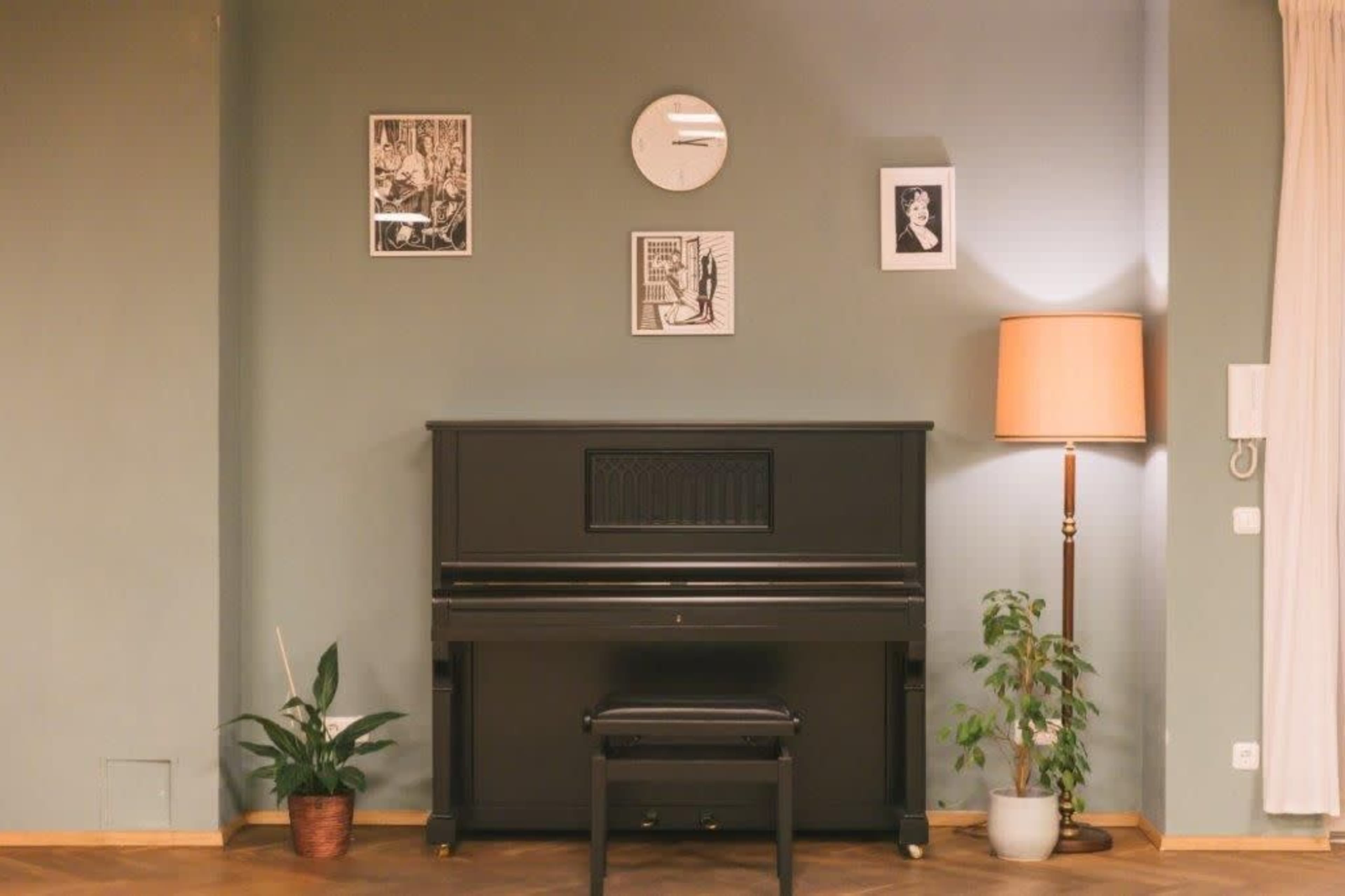 The image shows a room with a black upright piano, a lamp, a plant, a clock, and framed pictures on the wall.