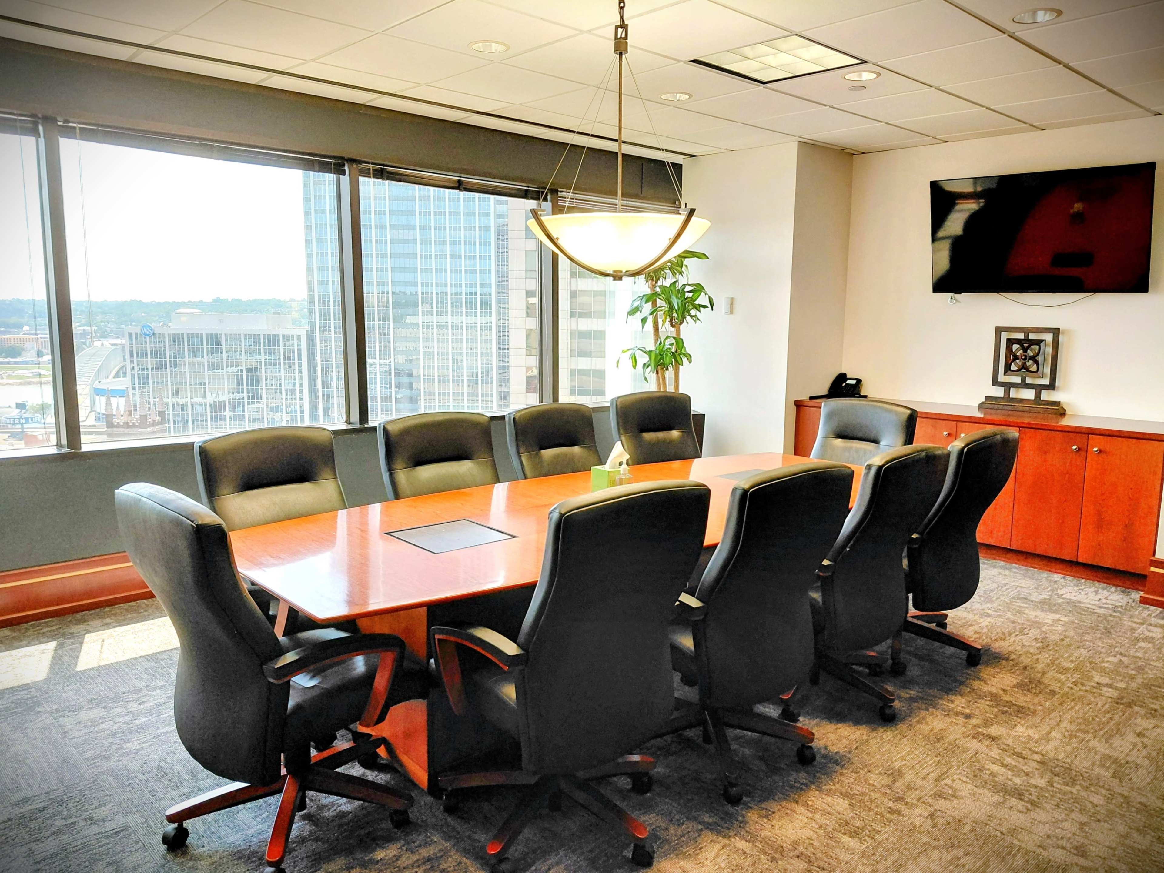 A spacious conference room features a large wooden table surrounded by ergonomic black chairs, with large windows providing a city view.