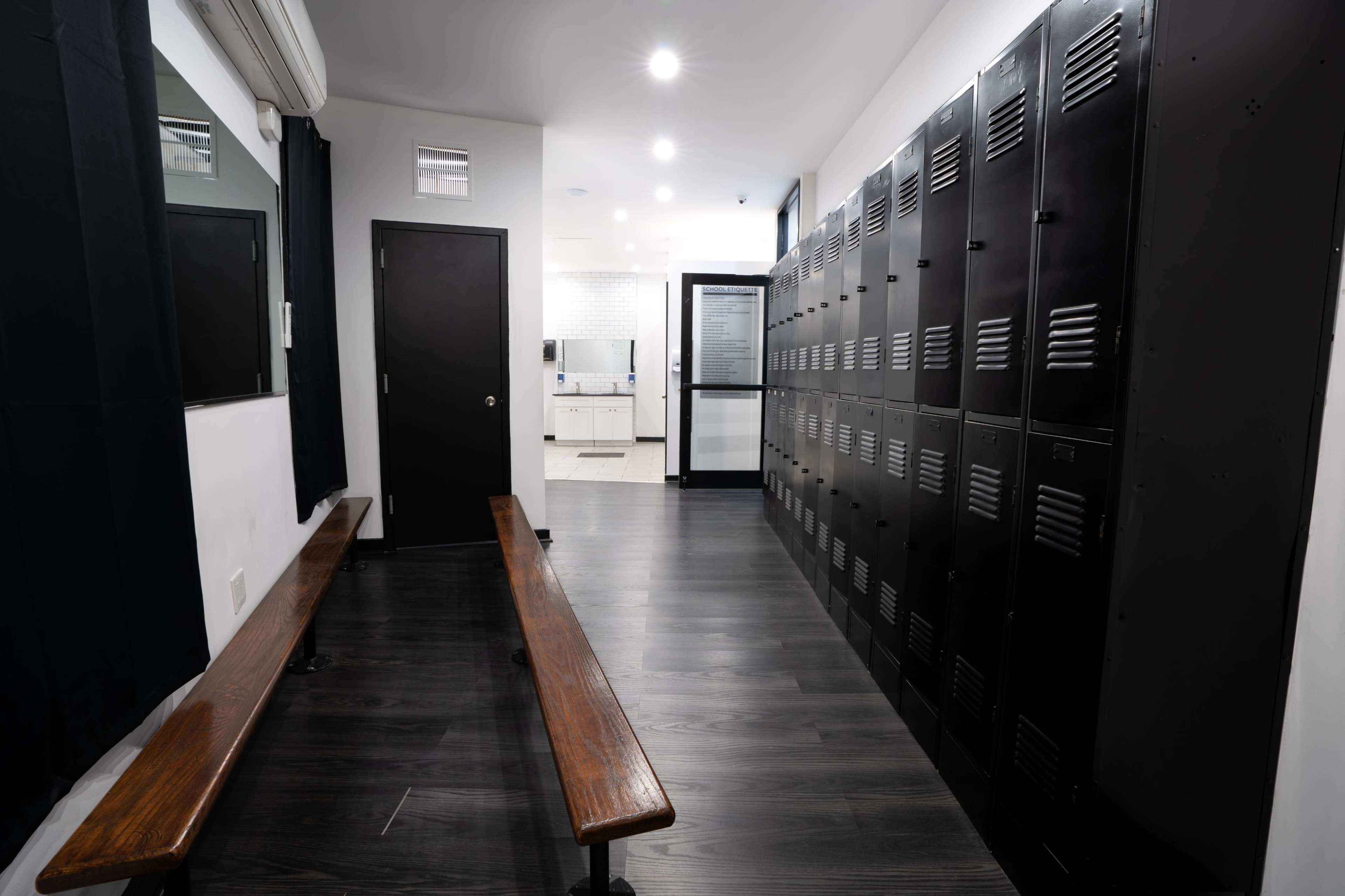 A hallway with two wooden benches and a row of black lockers against one wall, leading to a doorway at the far end.