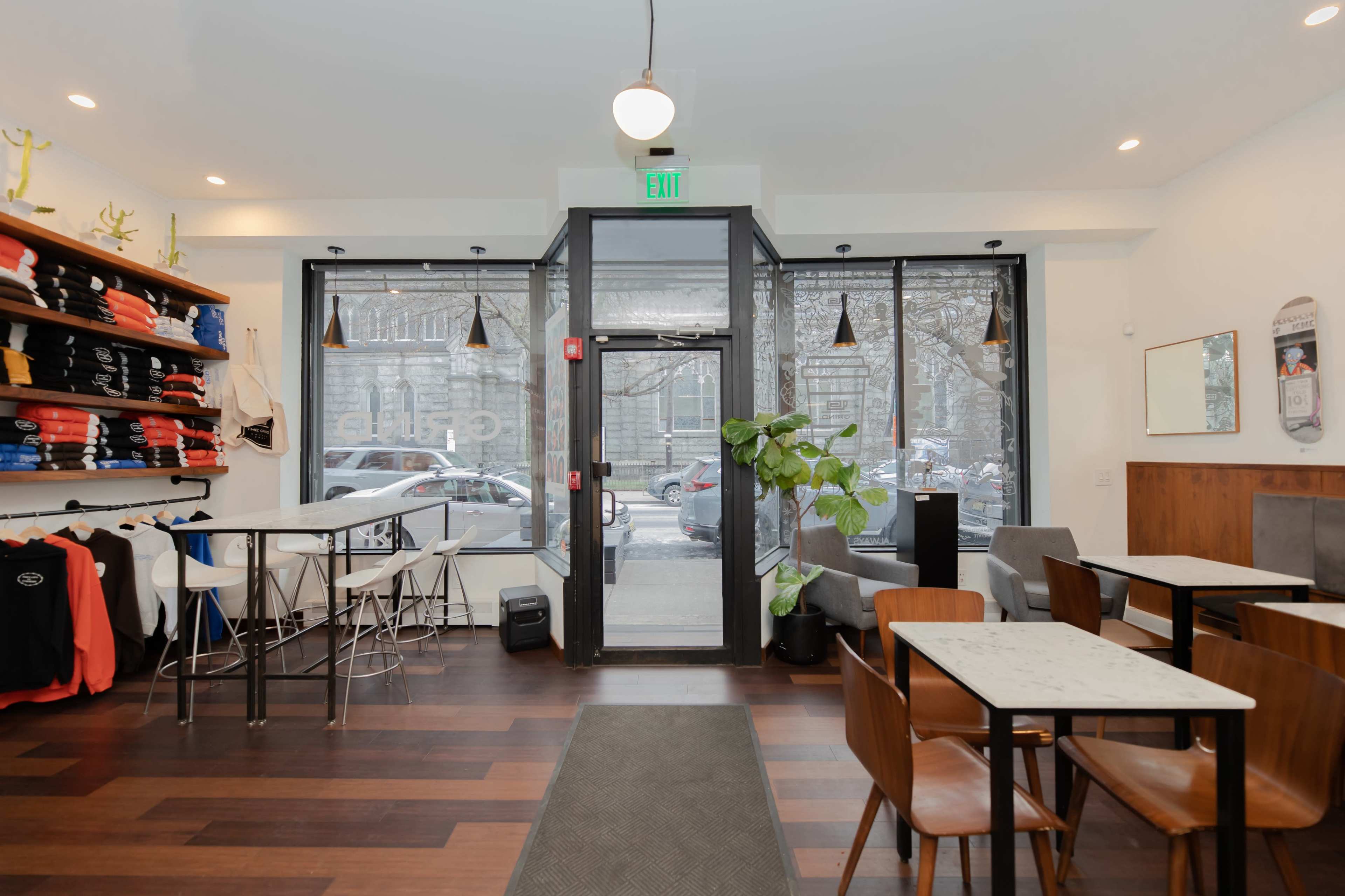 A modern café interior with tables, chairs, and a clothing display along one wall, featuring large windows that open to the street outside.