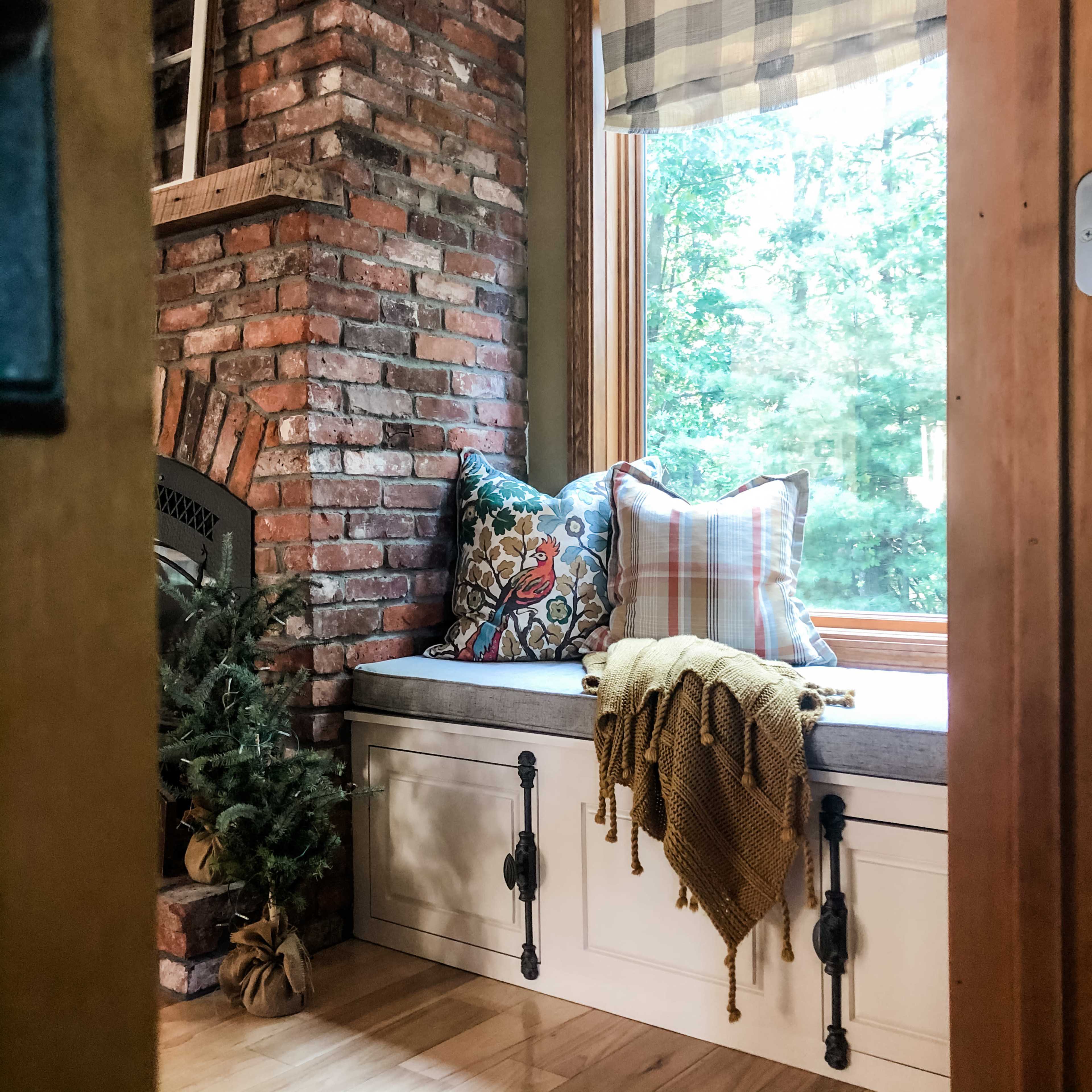 A cozy window nook with a cushioned bench, decorative pillows, and a throw blanket, adjacent to a brick fireplace and a window overlooking greenery.
