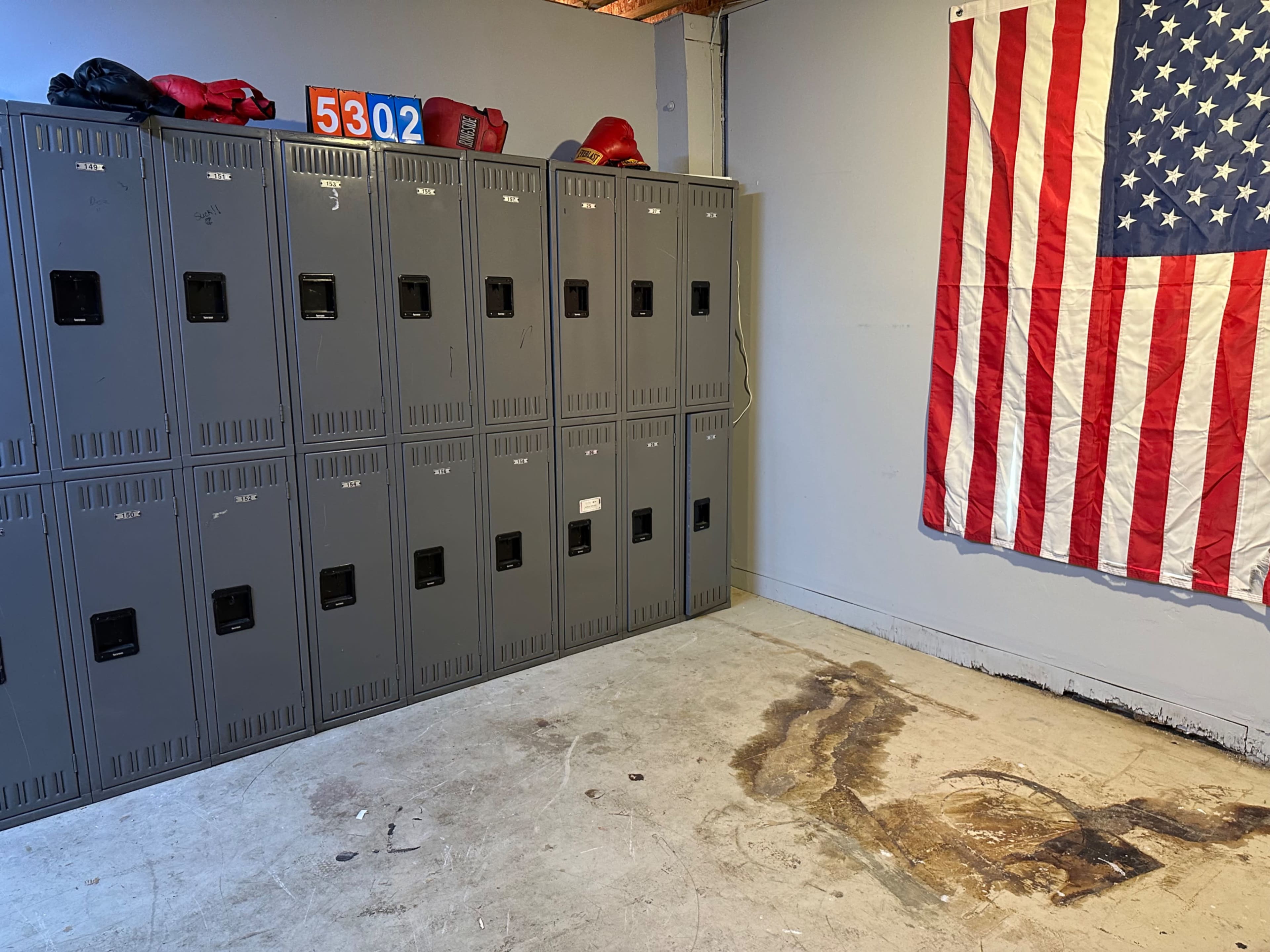 A room with gray lockers along one wall and an American flag hanging on another wall, with a stained floor in front of the lockers.