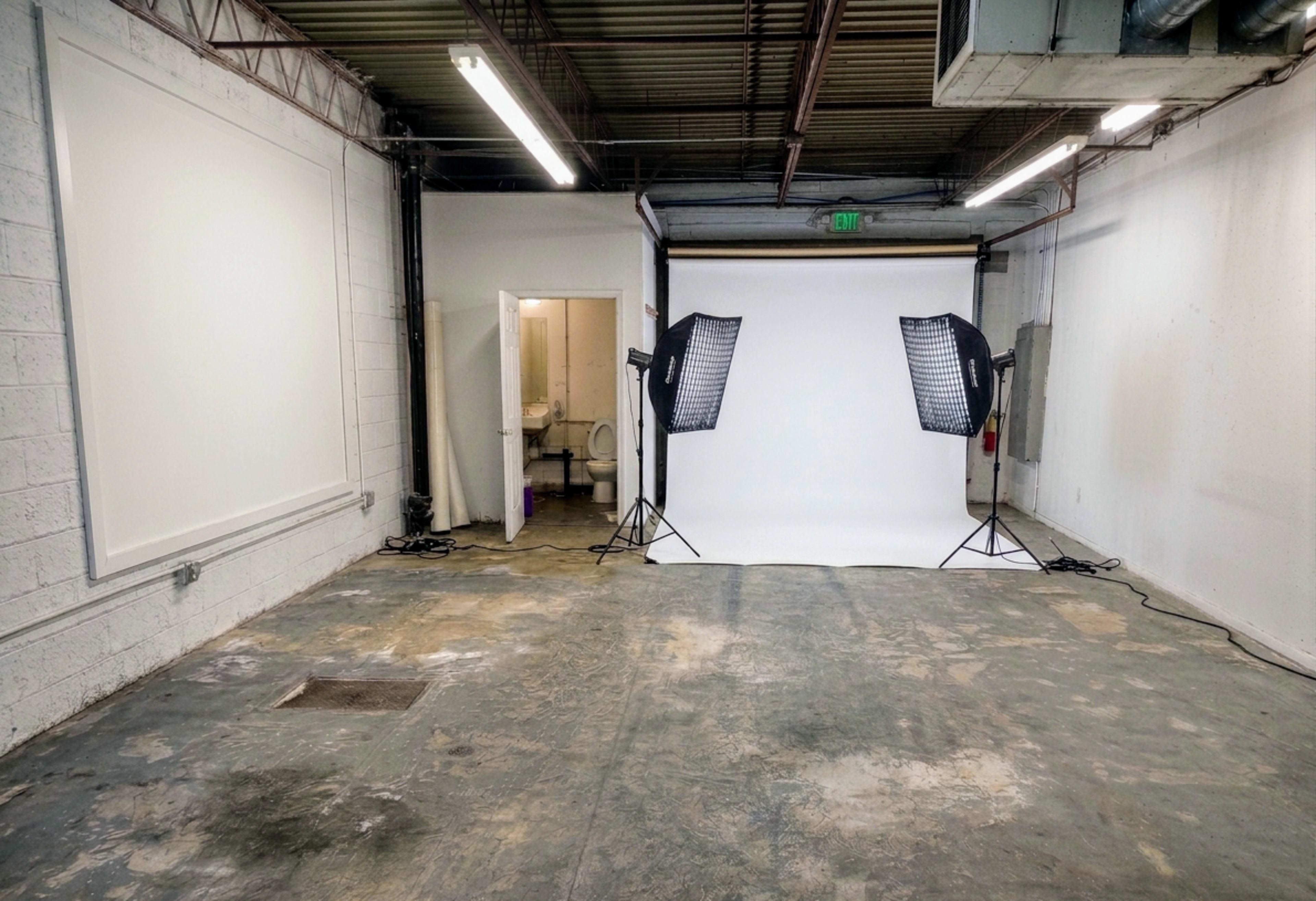 The image shows an empty studio space with a white backdrop, two lighting setups, and a doorway leading to a restroom.