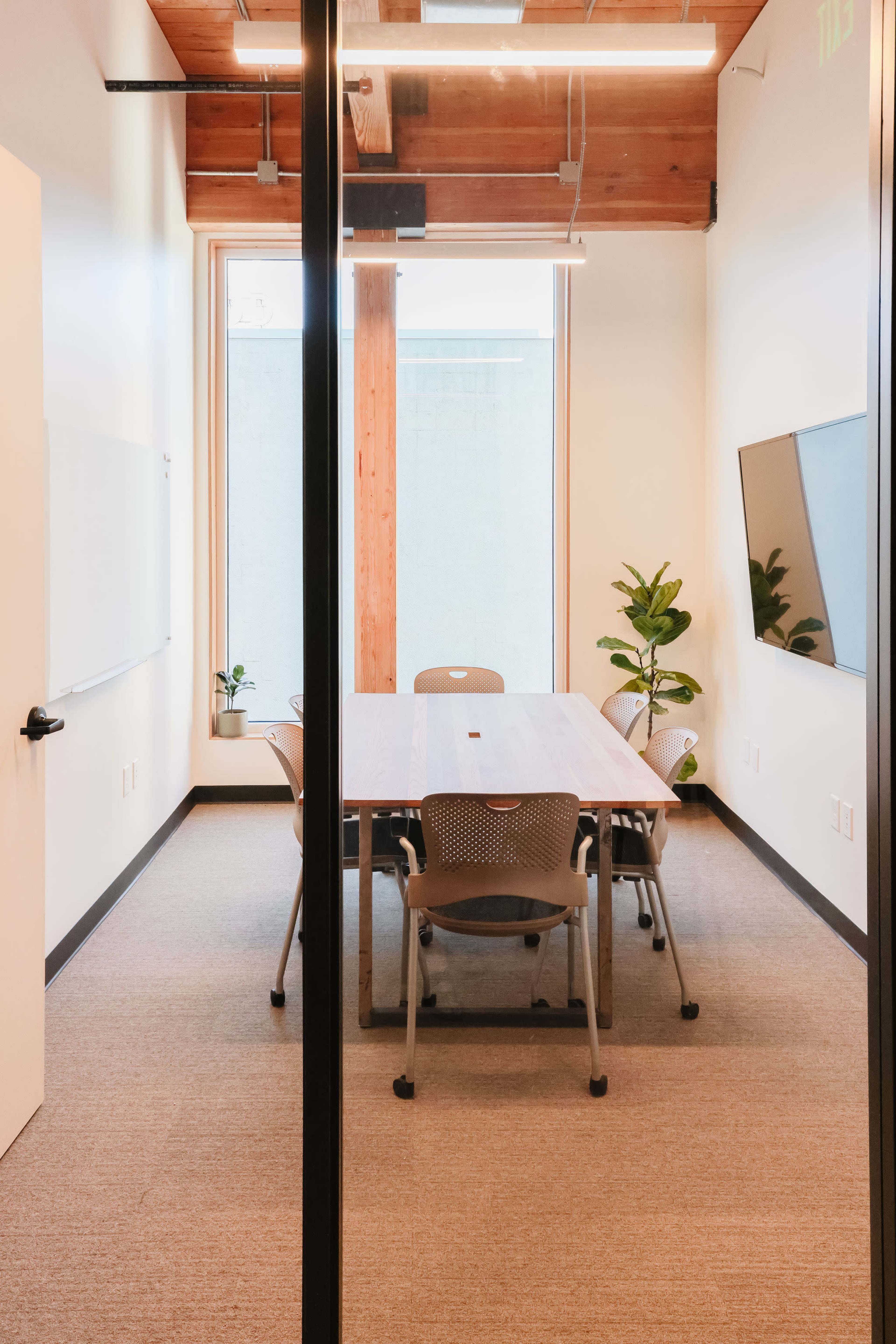 A small conference room features a wooden table surrounded by chairs, with a wall-mounted screen and plants near the windows.