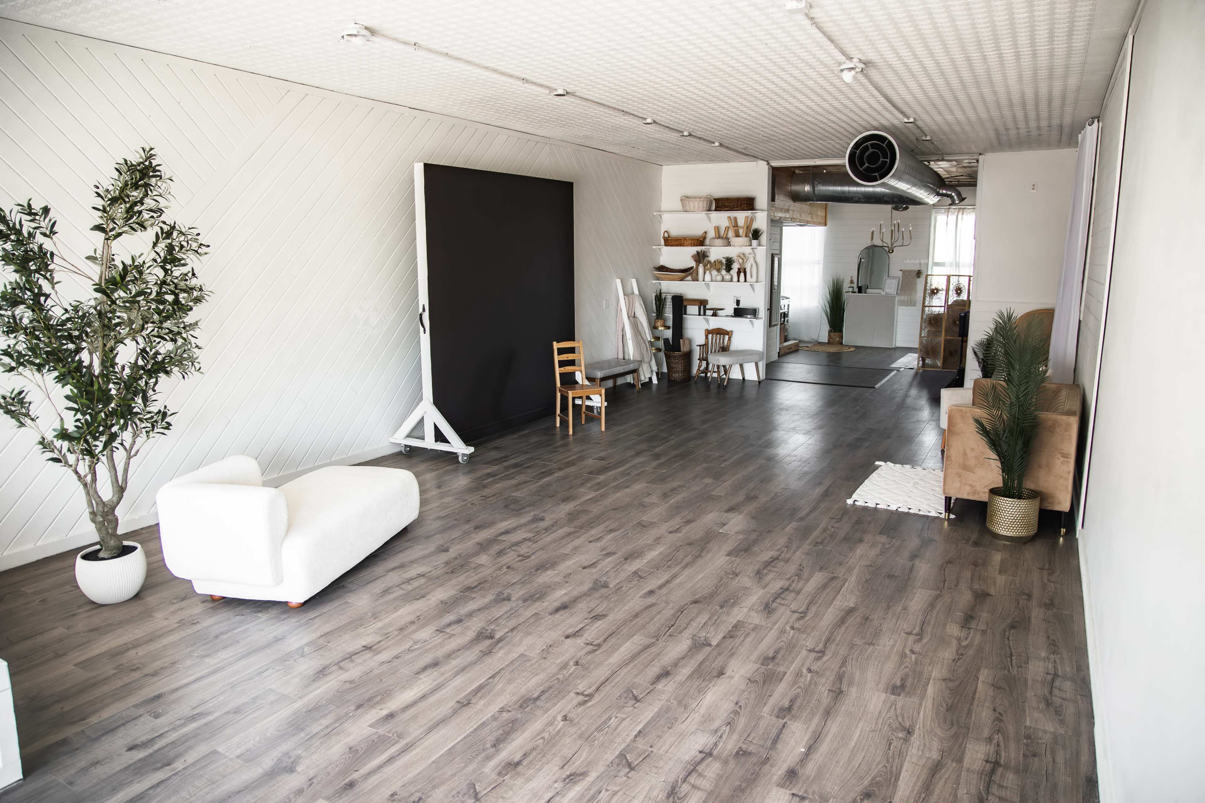 The image shows a spacious, minimally furnished room with wood flooring, featuring a large black backdrop on a stand, a white modern sofa, a brown chair, and several shelves with decorative items.