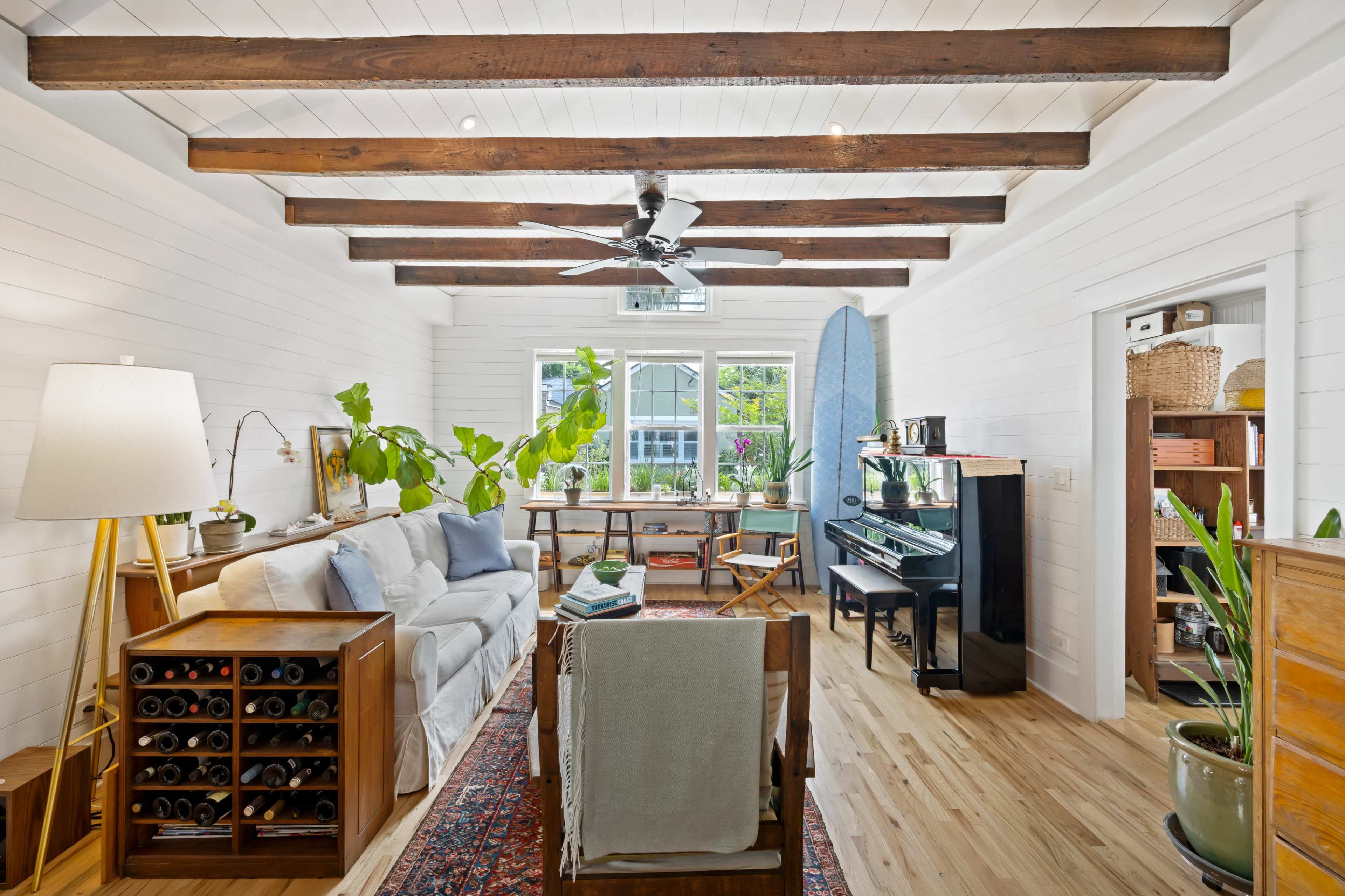 A bright living room features wooden beams, a ceiling fan, a piano, and a couch with plants and a wine rack nearby.