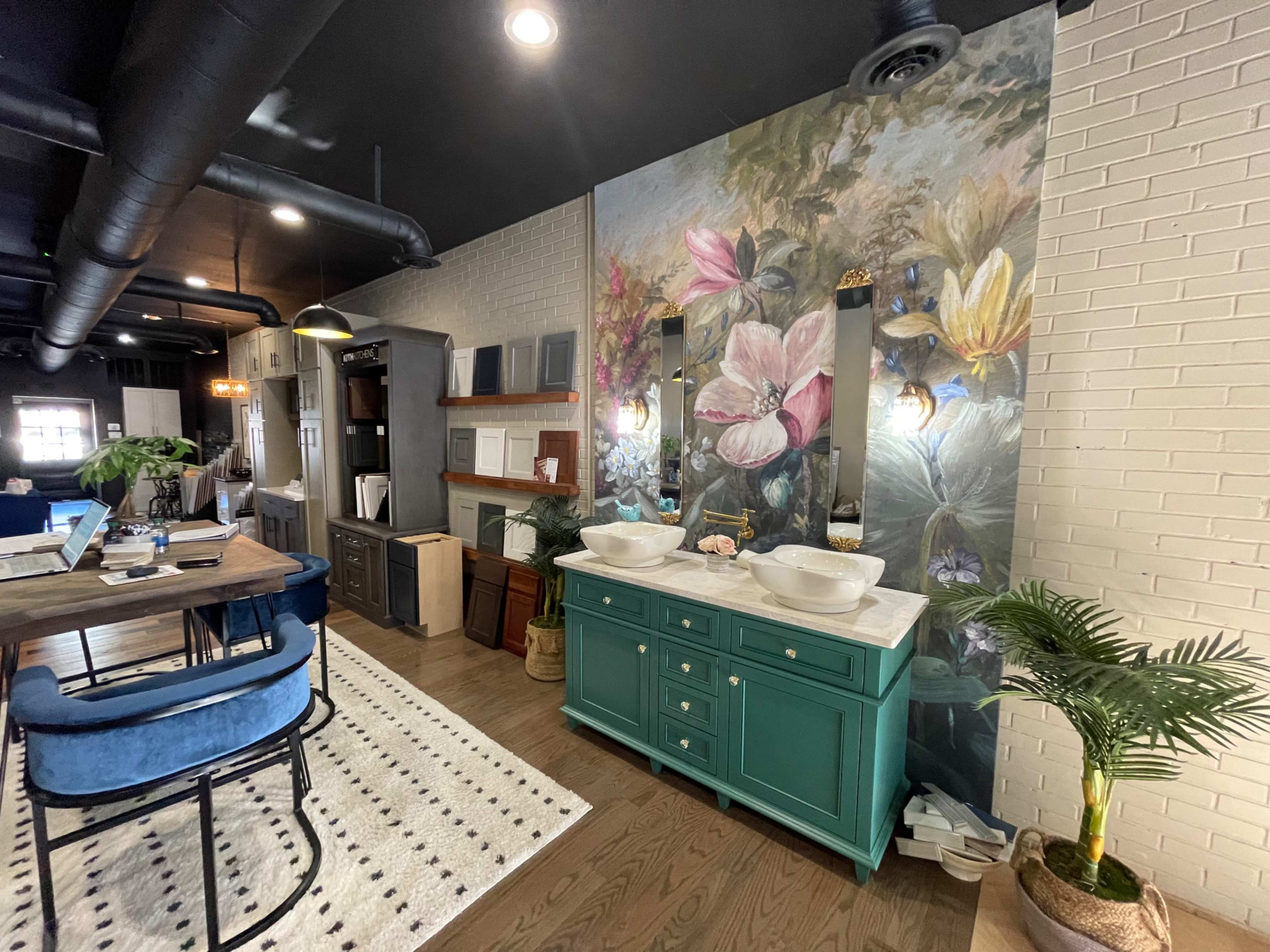 A stylish interior featuring a green vanity with two sinks, a floral mural on the wall, wooden shelves, and a desk in a well-lit space.