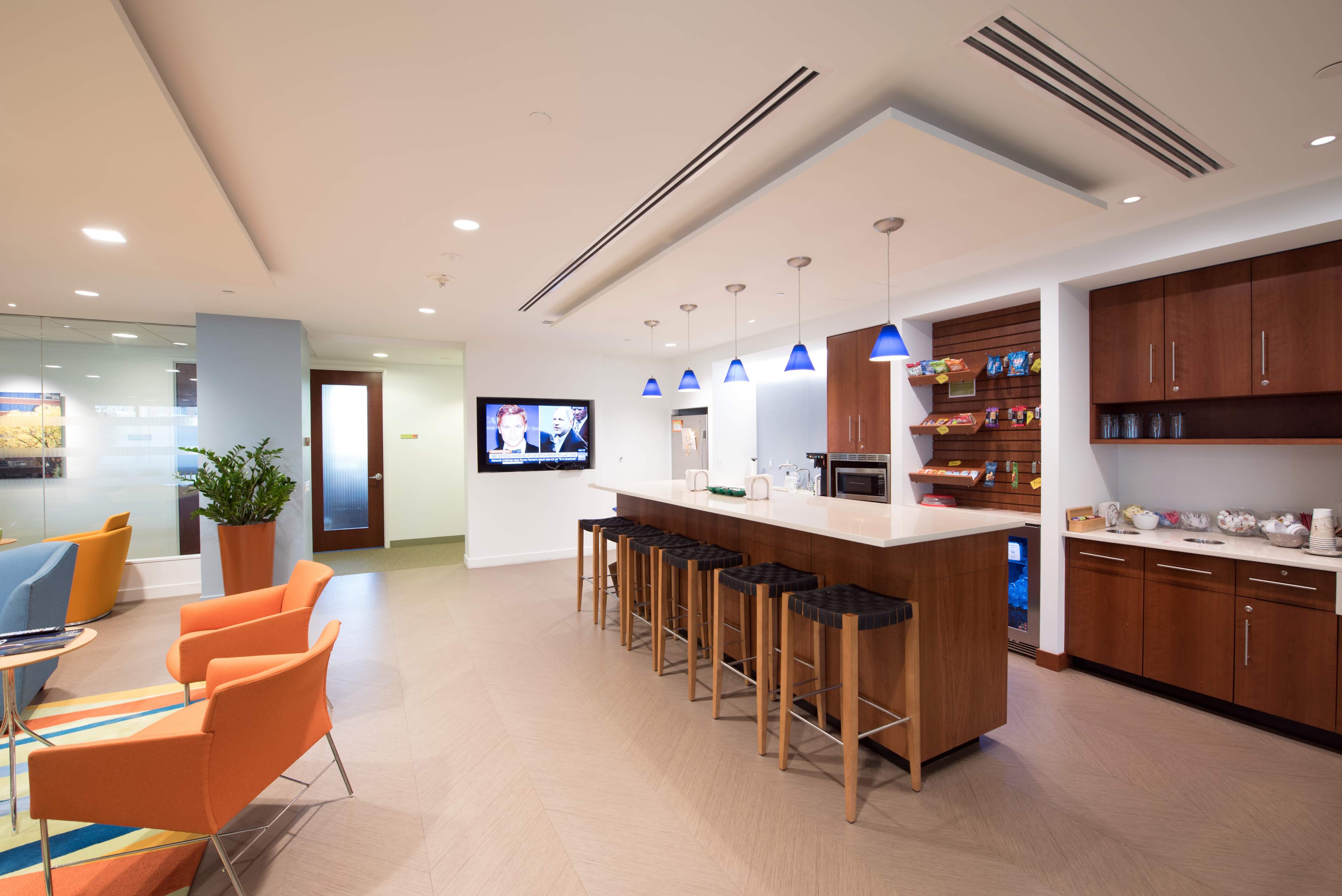 A modern kitchen and lounge area with a bar seating, a television on the wall, and various snacks displayed on shelves.