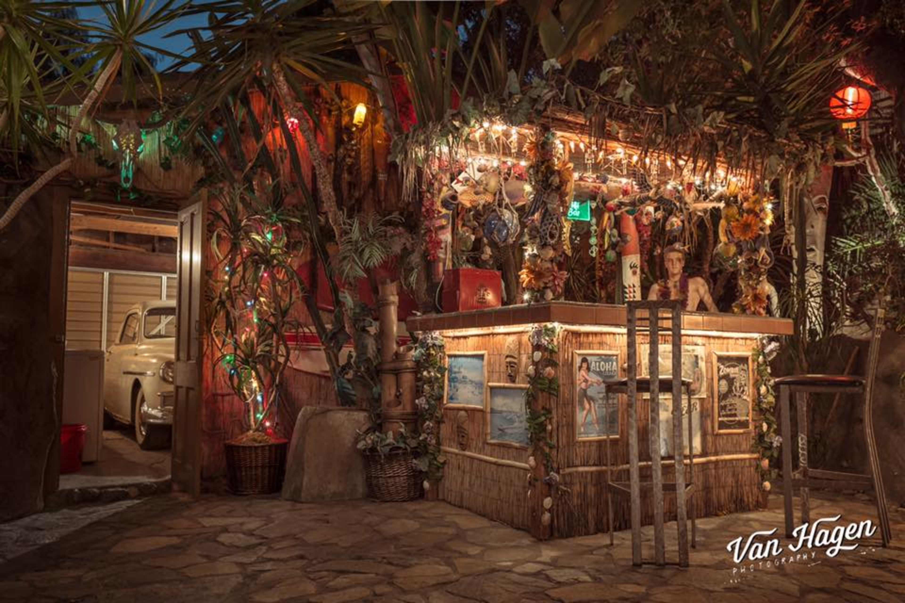 A tropical-themed bar with twinkling lights and decorations surrounded by lush foliage, positioned next to a vintage car partially visible in an open garage.