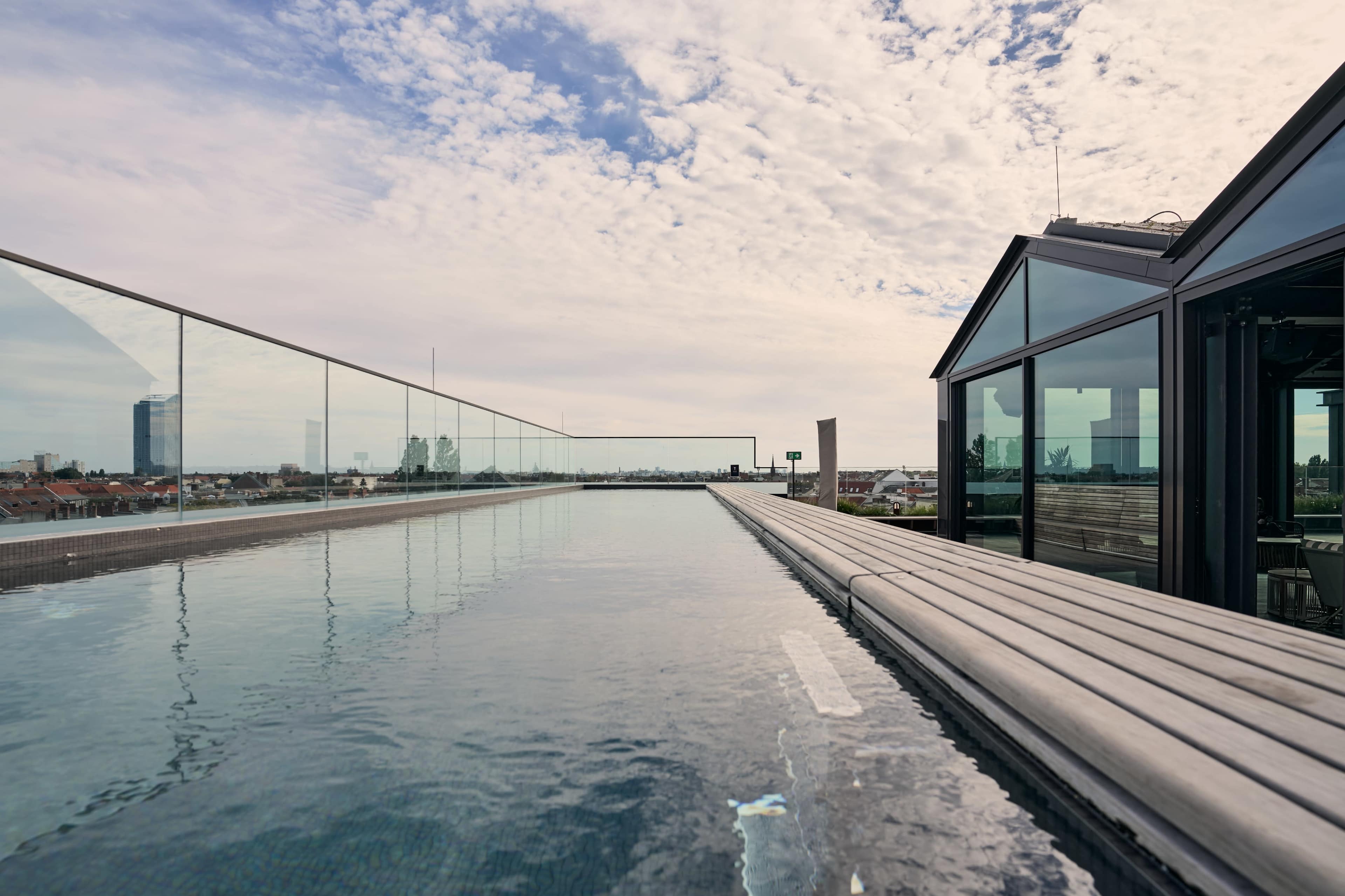 The image shows a modern rooftop pool with a glass railing, reflecting the sky and surrounding buildings.