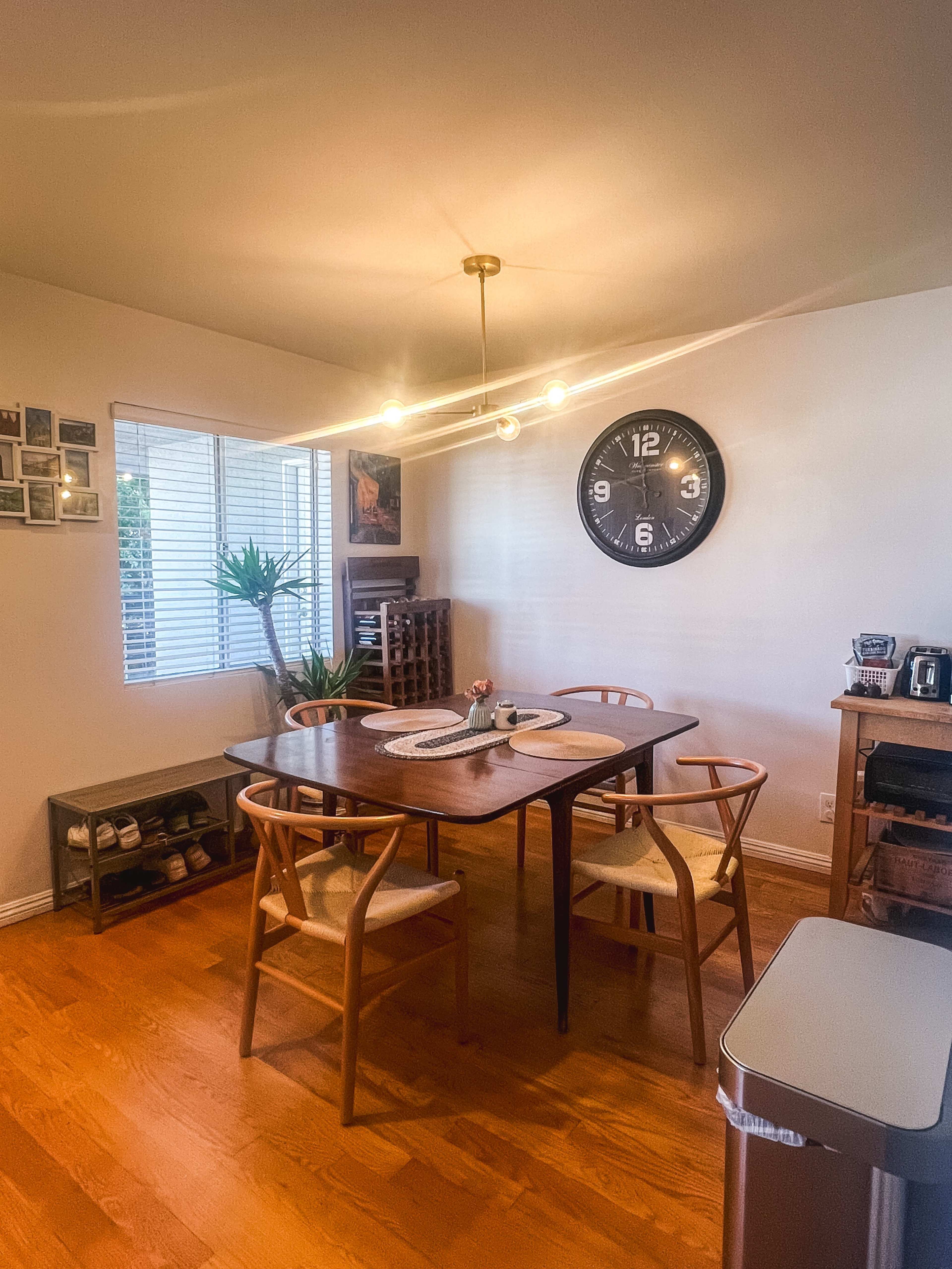 A dining area features a wooden table with six chairs, a wall clock, and a window providing natural light.