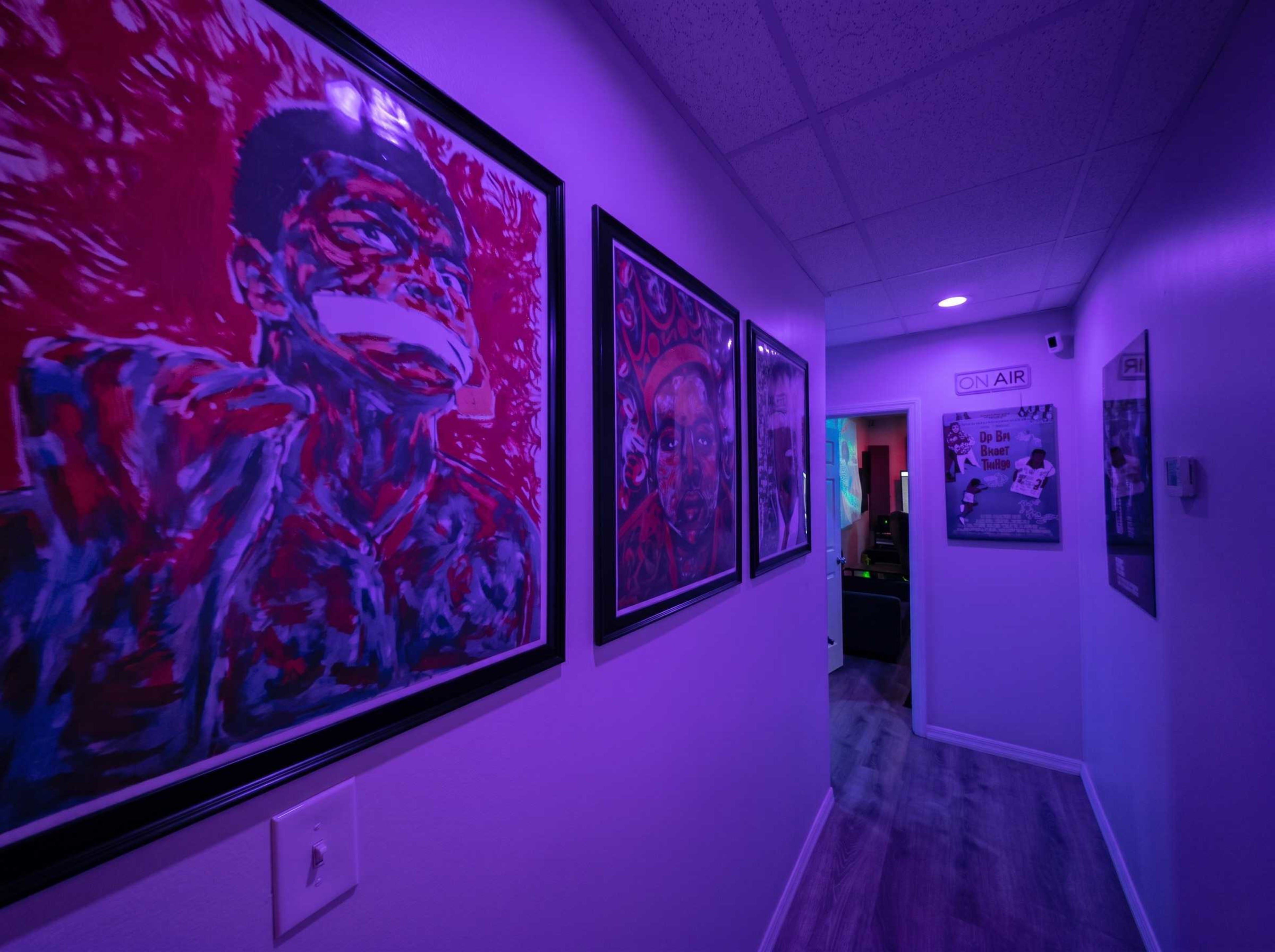 A narrow hallway is lined with vibrant framed artwork, illuminated by purple lighting.