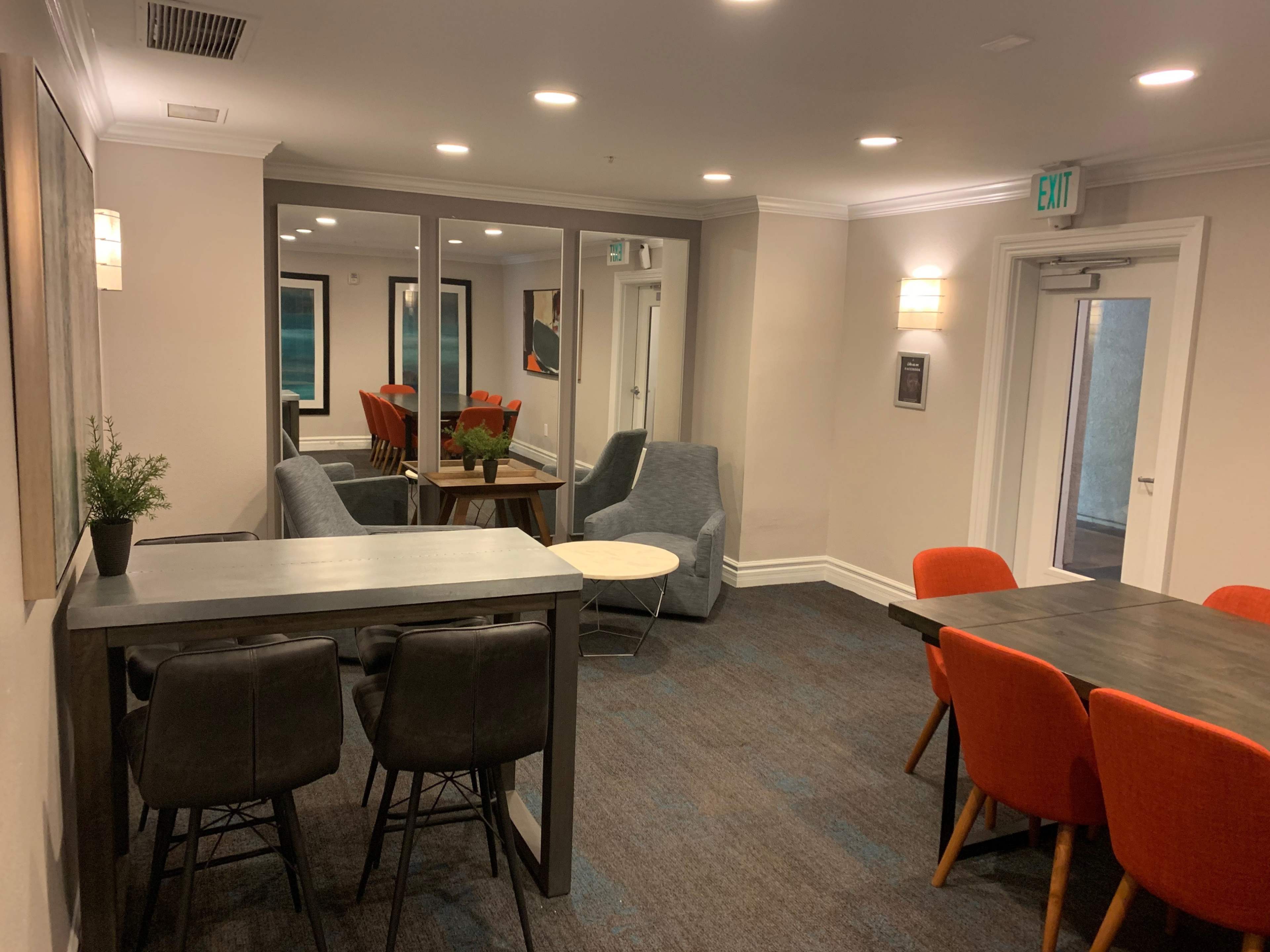The image shows a modern lounge area featuring gray seating, a small round table, and a dining space with orange chairs.