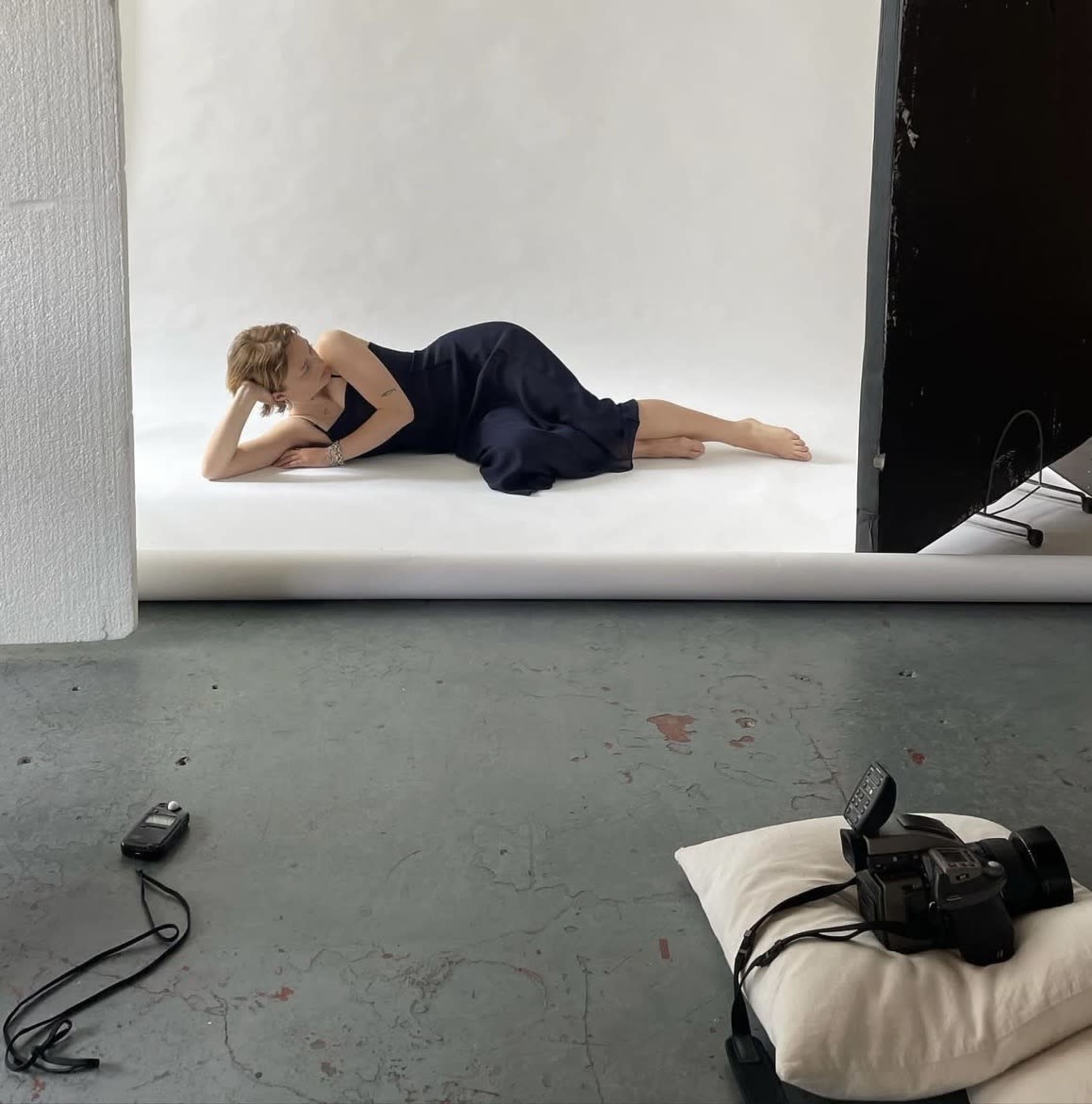 A model in a dark dress lies on a white backdrop in a photography studio, while a camera rests on a pillow nearby.