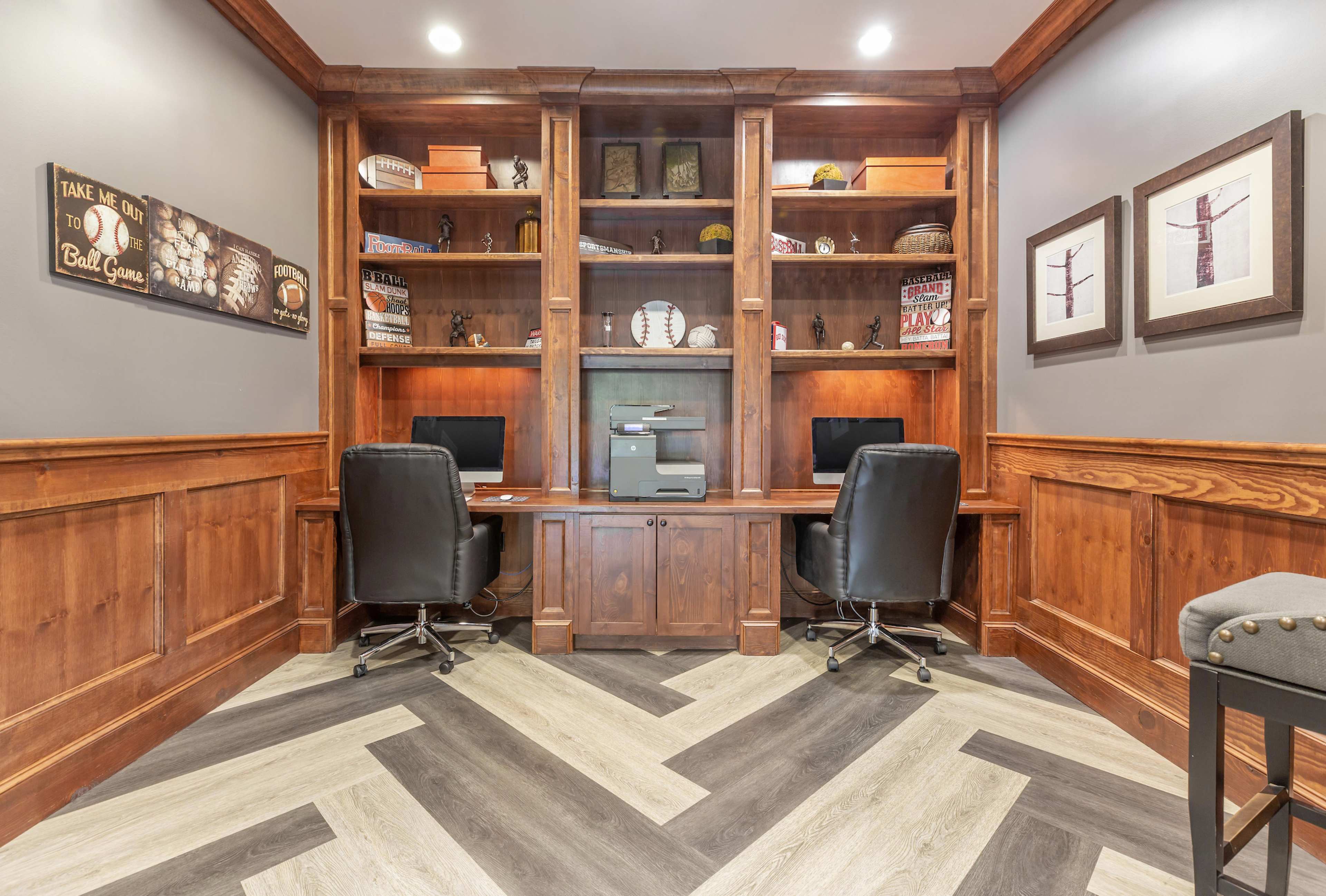 A dual workspace with two black office chairs, a printer, and built-in wooden shelves displaying various collectibles and decor.