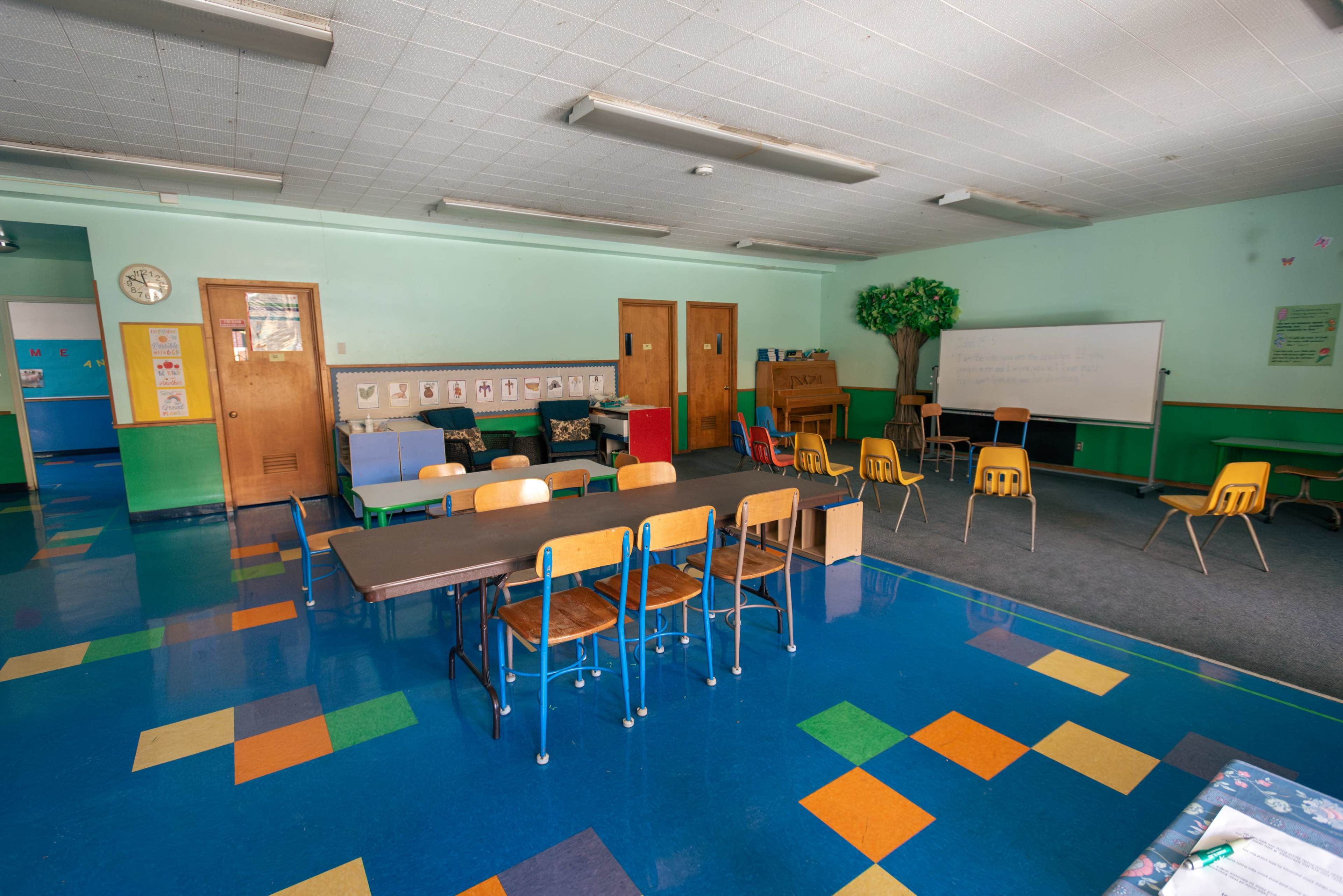 A classroom is arranged with multiple tables and chairs, featuring a green wall, a large whiteboard, and a few decorative elements.