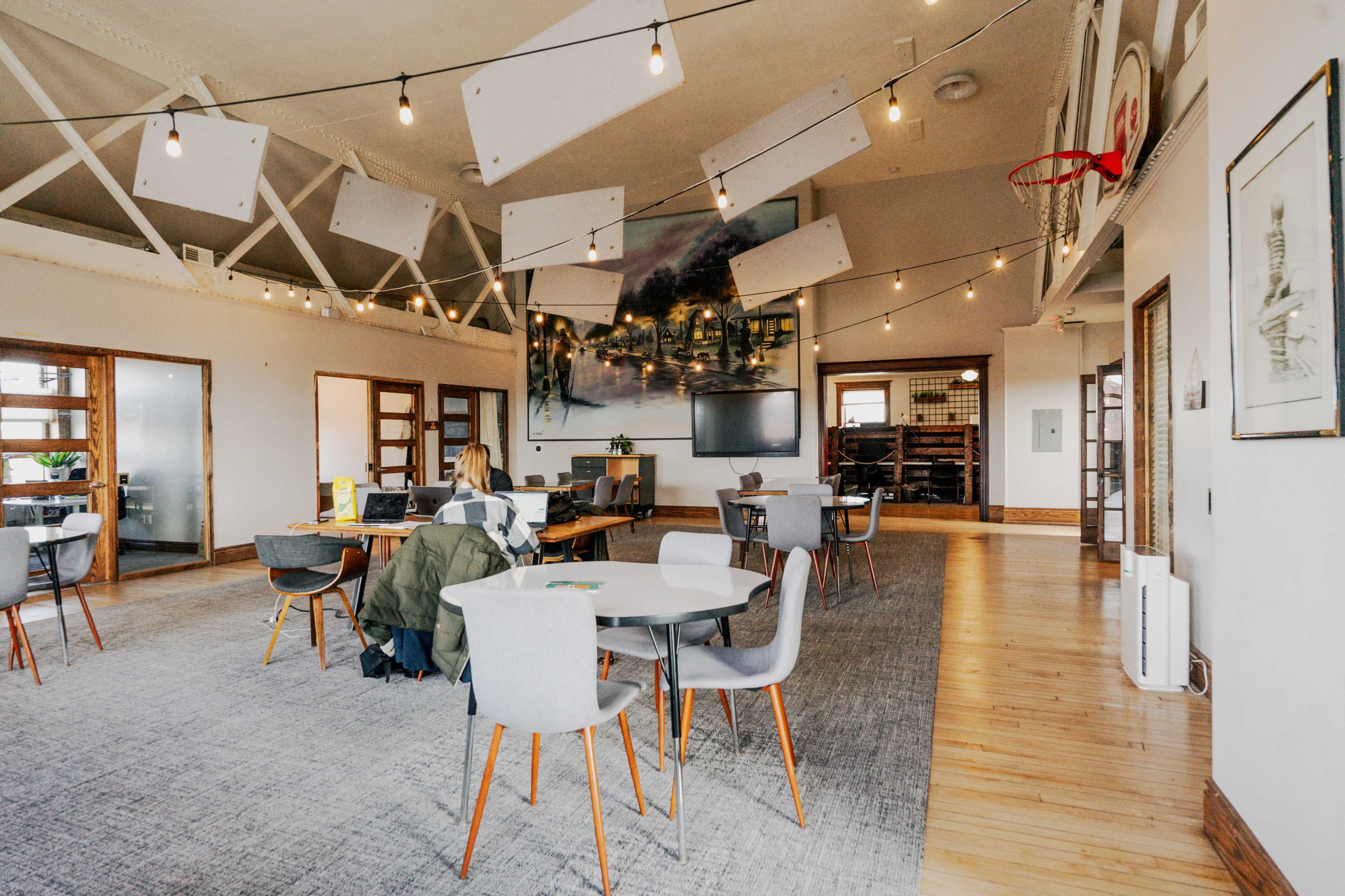 A spacious, modern office with several tables and chairs, overhead sound panels, and a large wall painting, occupied by a woman working at one of the tables.