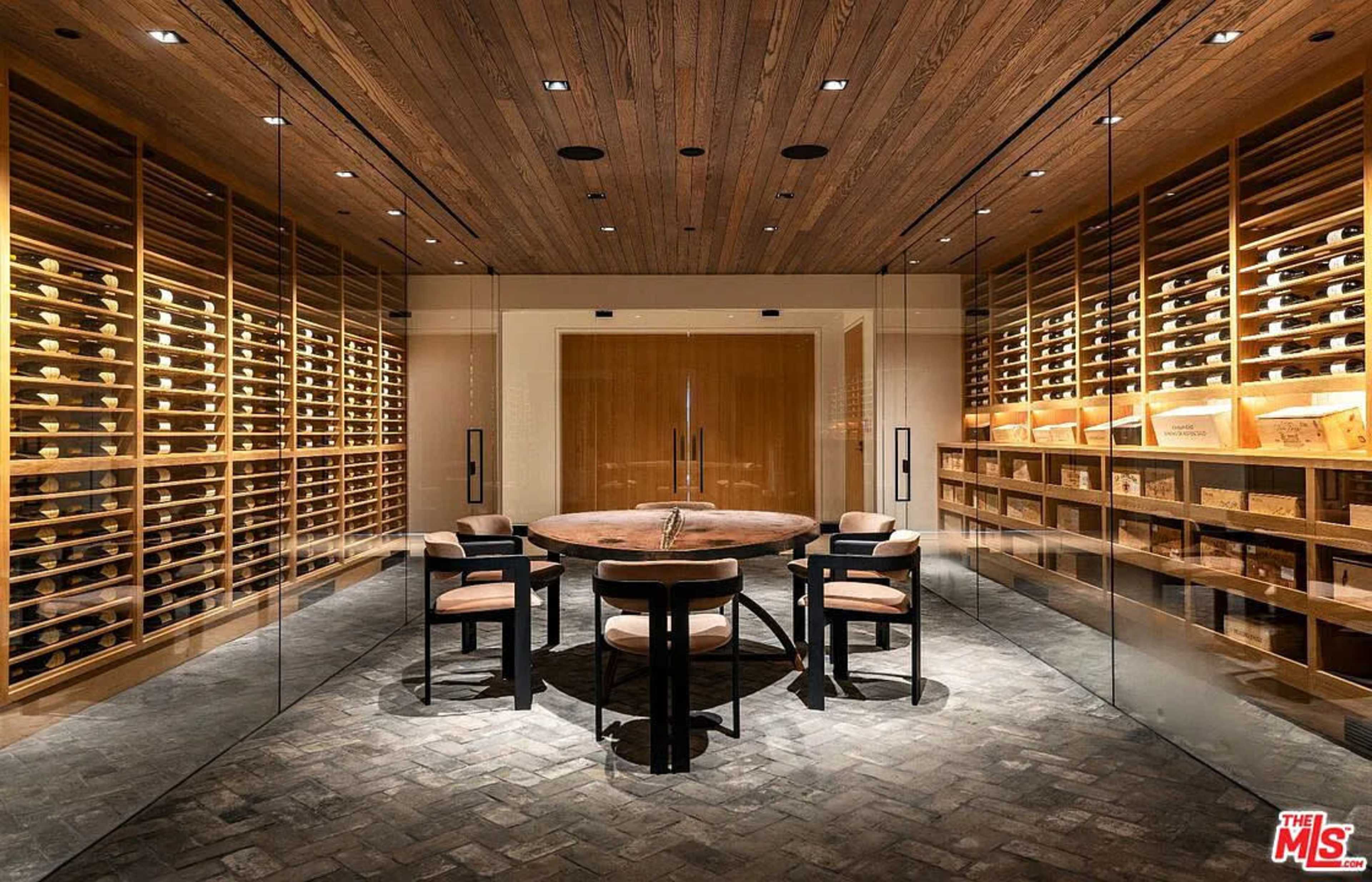 The image shows a modern wine cellar featuring a circular wooden table surrounded by four chairs, with glass walls and shelves filled with wine bottles.