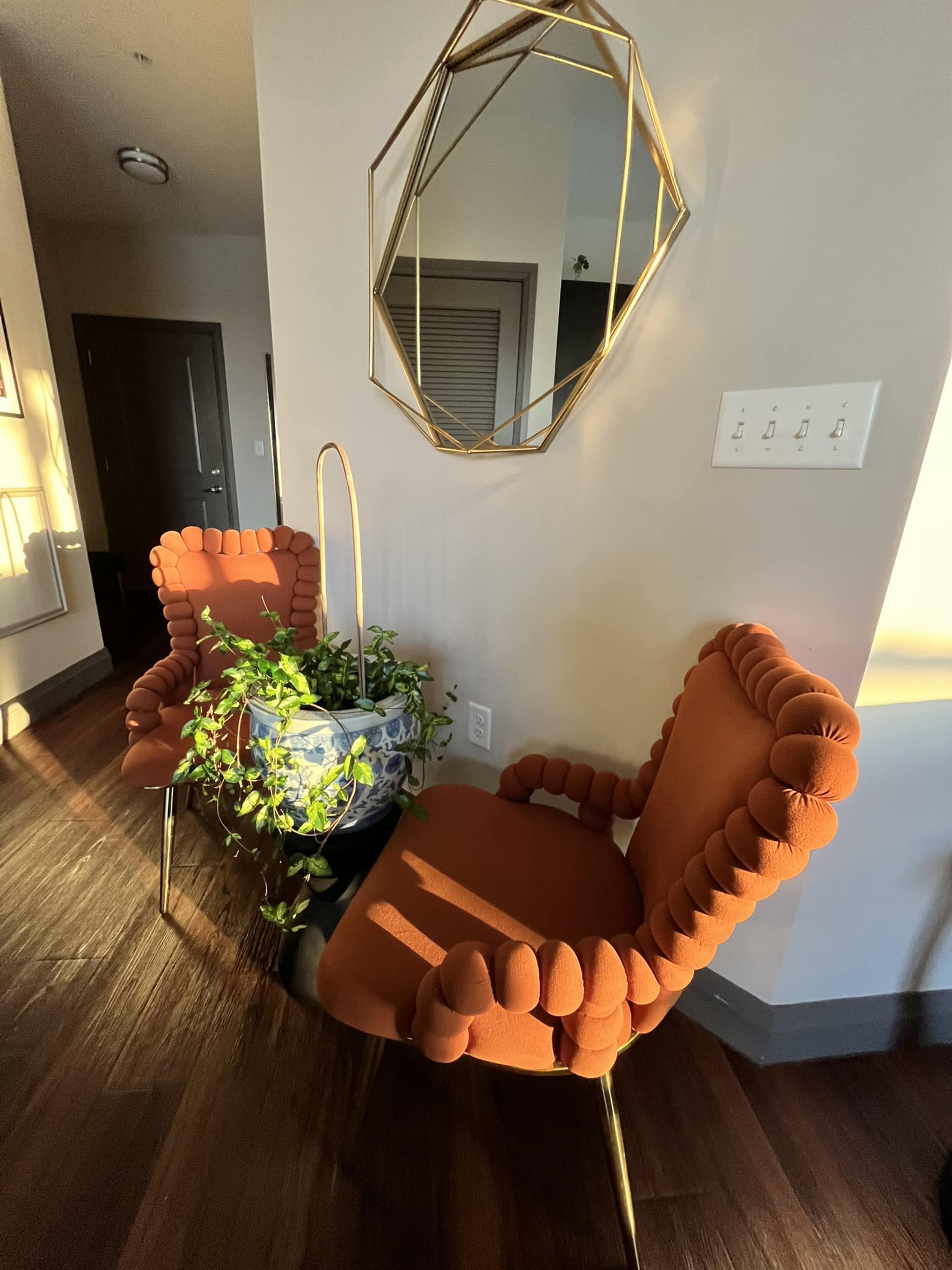 Two orange textured chairs flank a round table holding a potted plant beneath a geometric mirror on a wall with light streaming in.