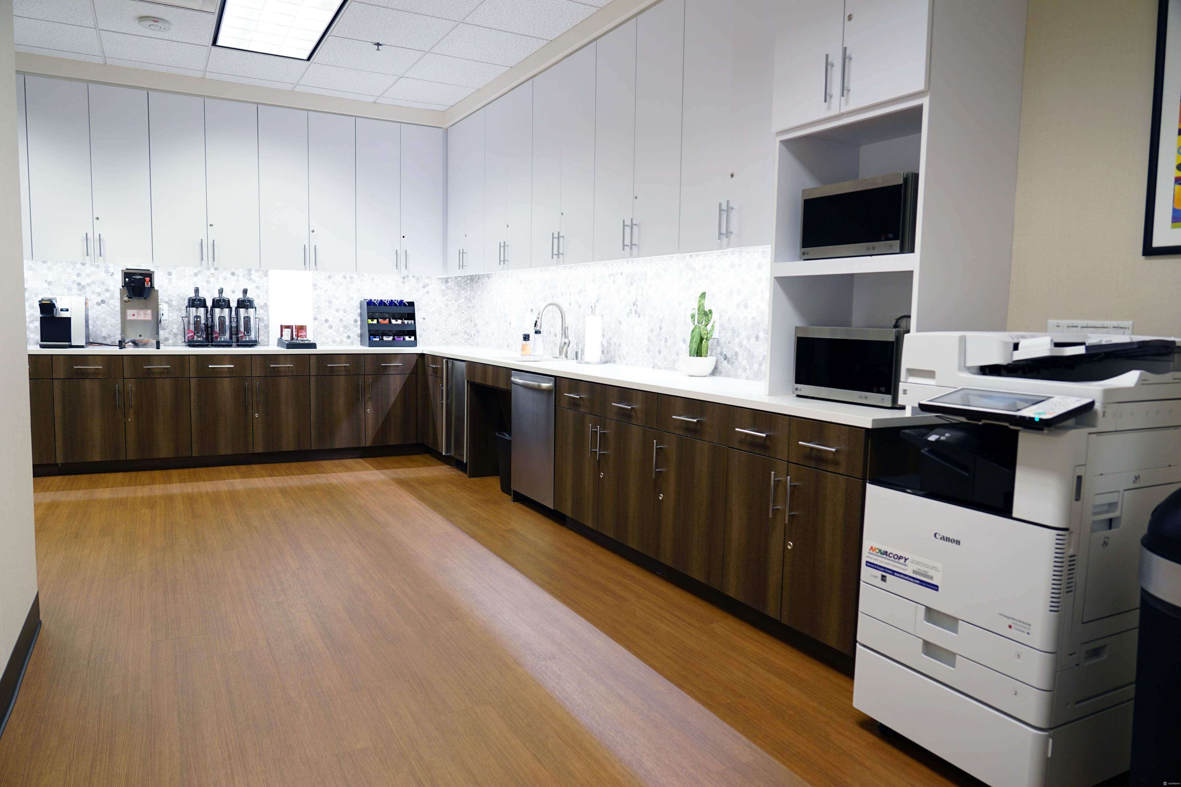 The image depicts a modern office kitchen area with wooden cabinets, a marble backsplash, a coffee machine, microwaves, a sink, and a printer.