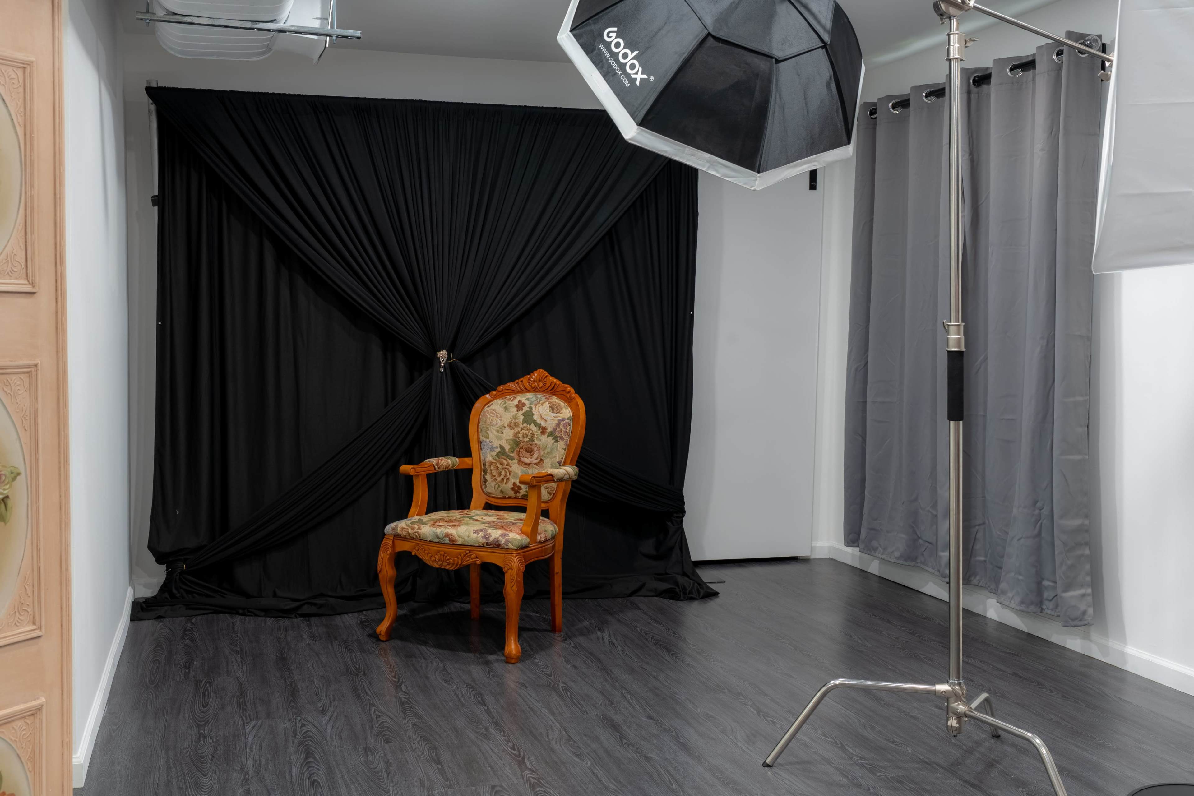 The image shows a photography studio featuring a decorated wooden chair in front of a black backdrop, with softbox lighting equipment to the side.
