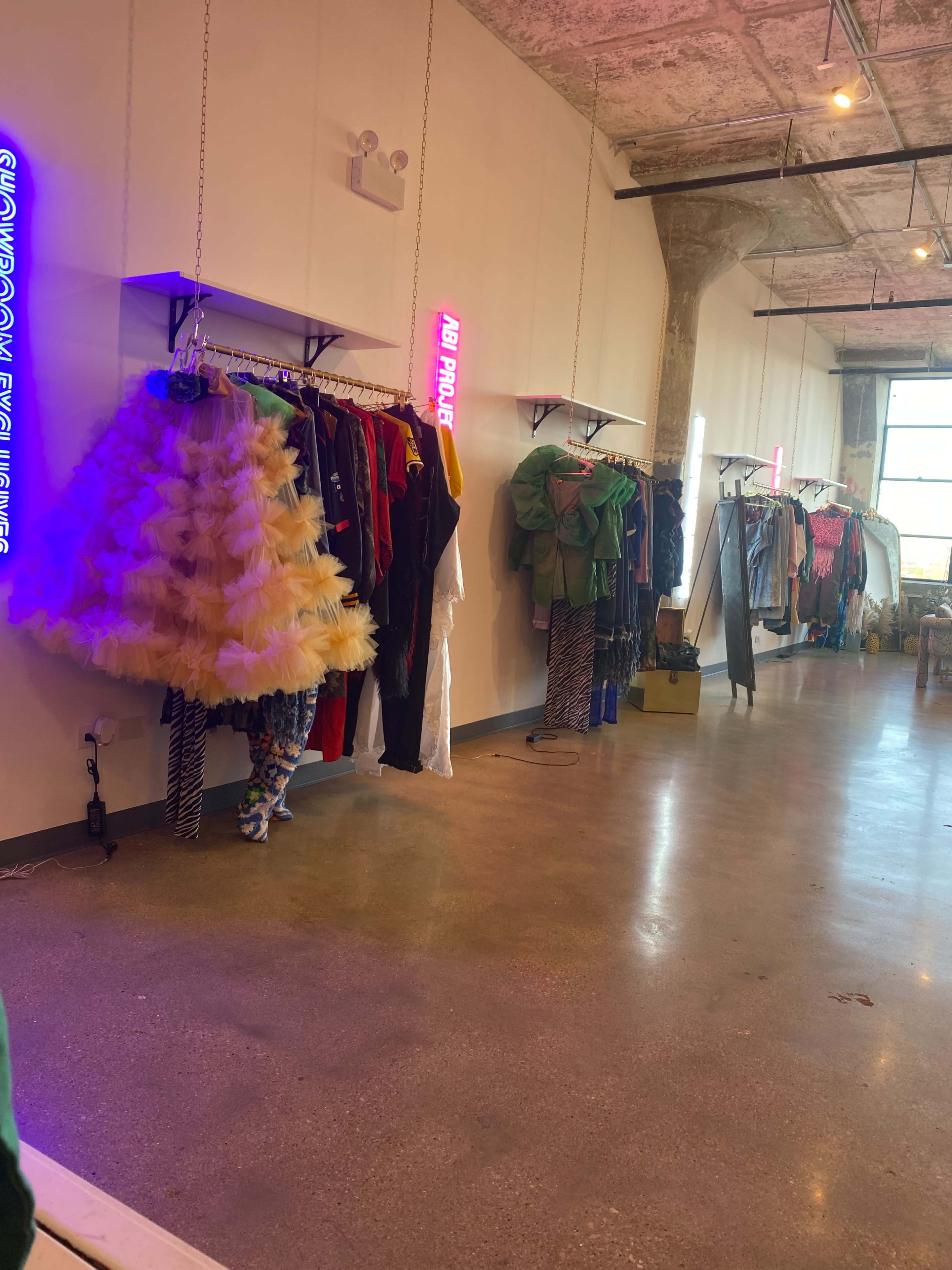 The image shows a clothing store interior with various garments displayed on hangers along the walls, and neon signs illuminating the space.
