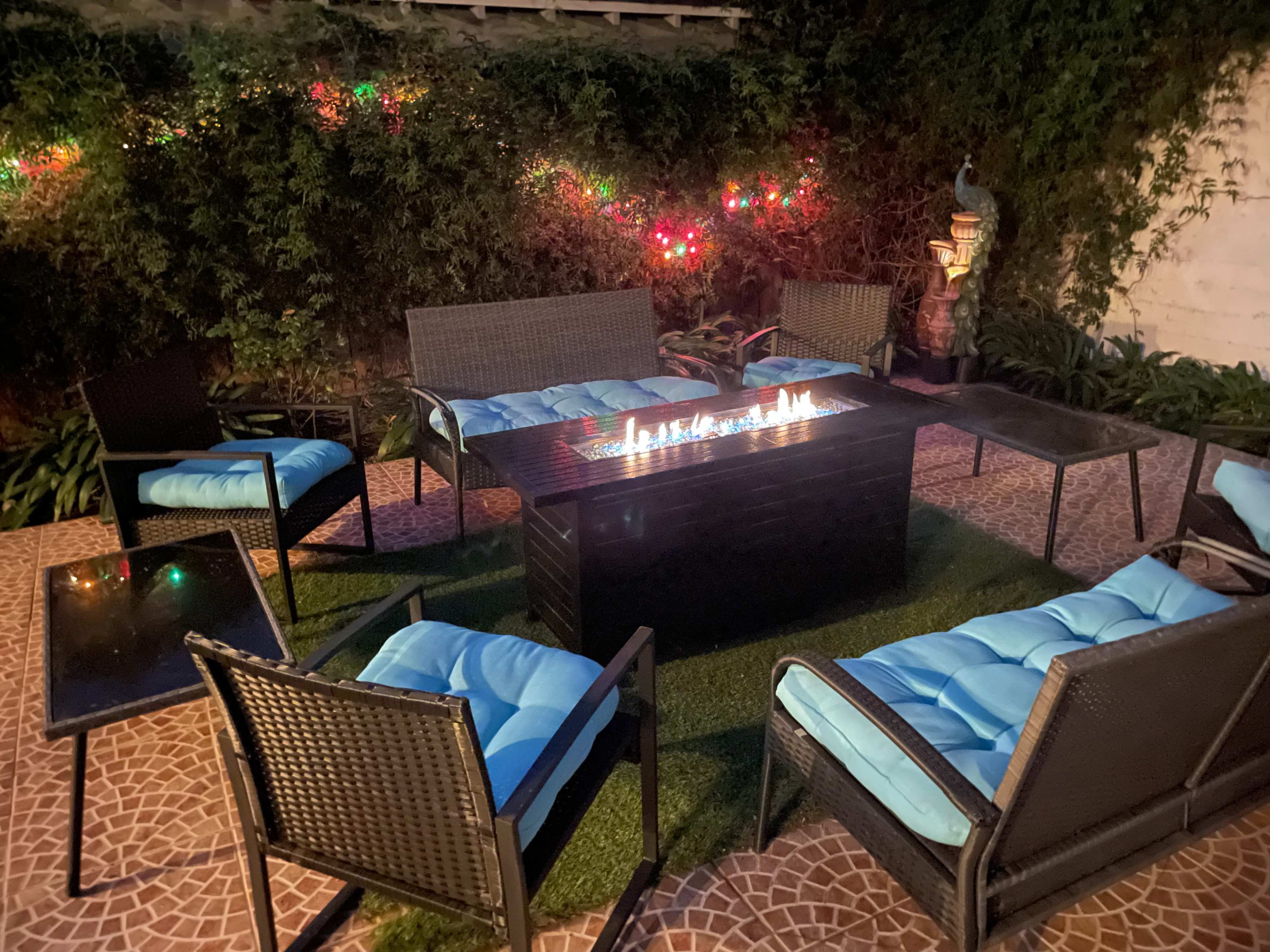 A patio setting features a rectangular fire pit surrounded by six chairs, with colorful string lights illuminating a green backdrop.