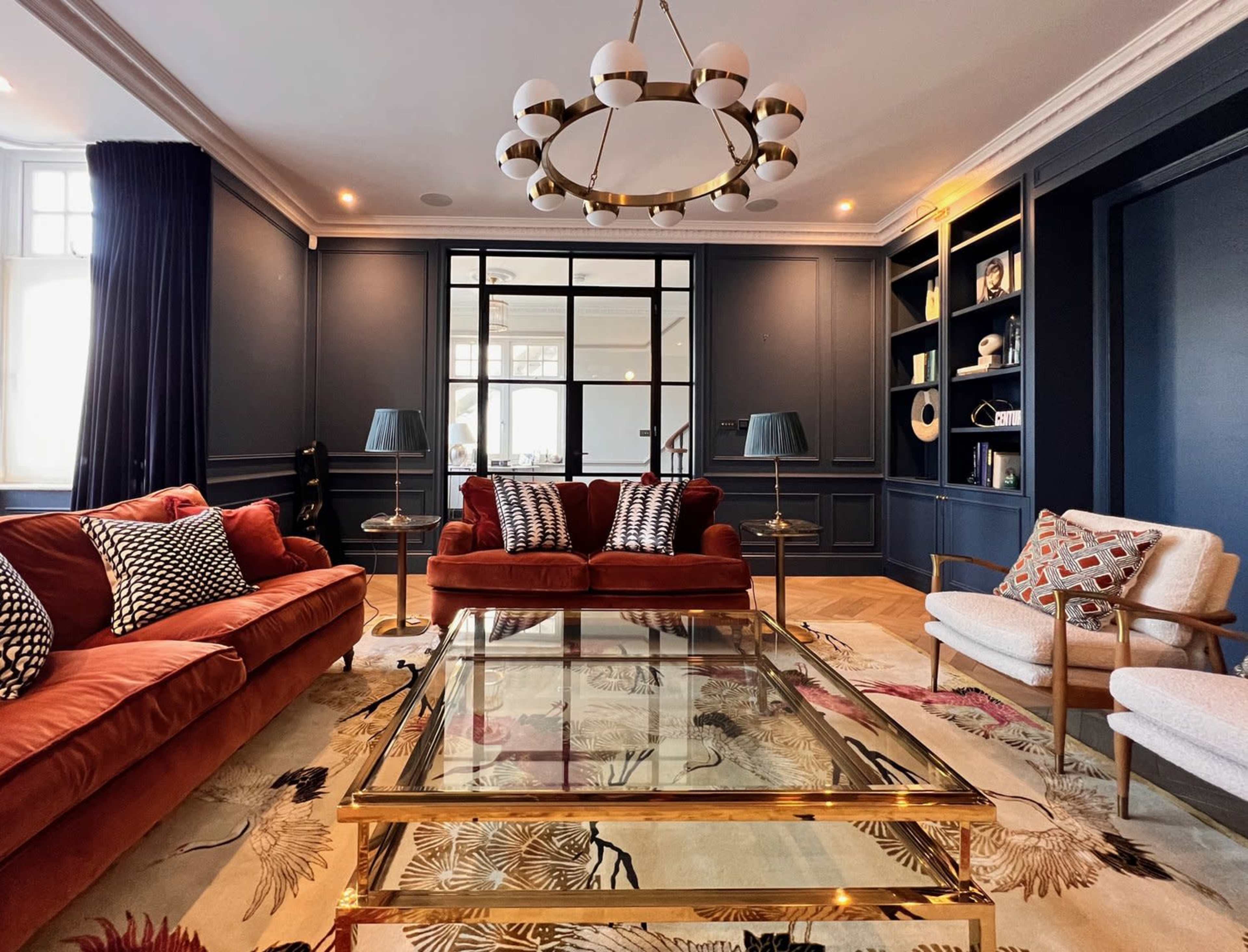 A stylish living room features a brown velvet sectional sofa, a glass coffee table, and dark blue walls embellished with built-in shelves and modern lighting fixtures.