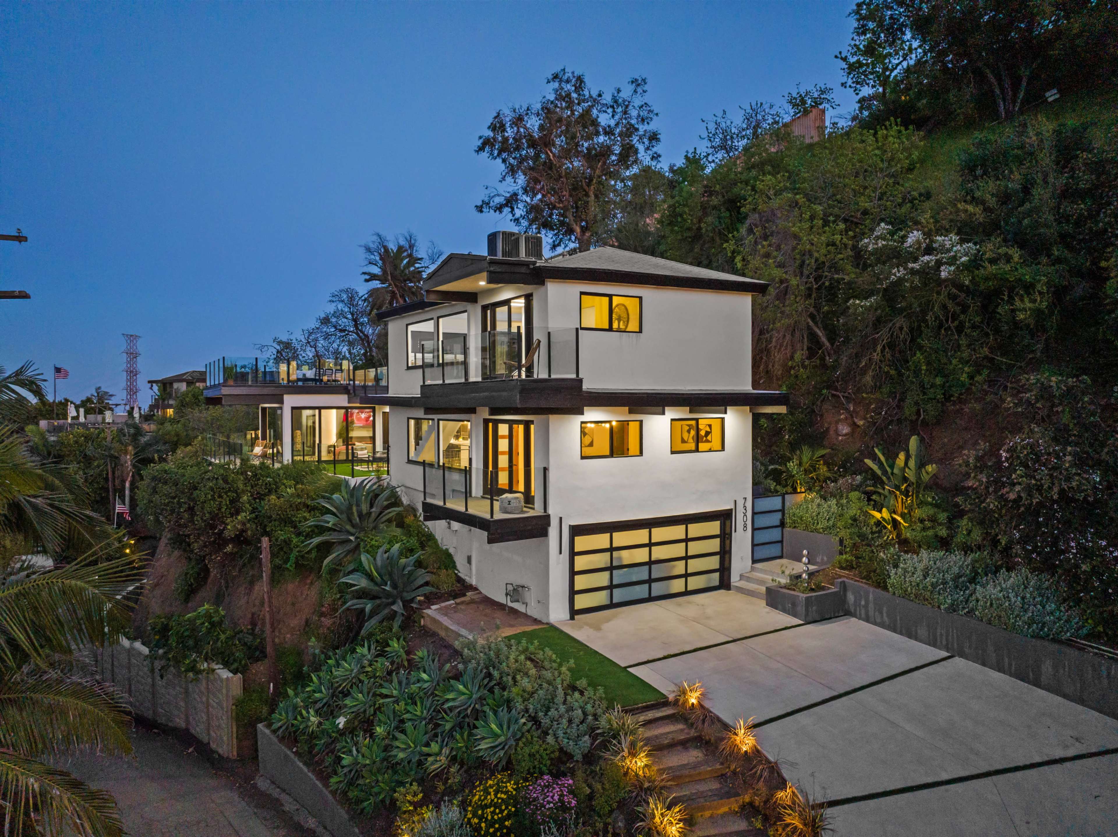 A modern two-story white house with a garage and landscaped gardens is situated on a hillside, illuminated at dusk.