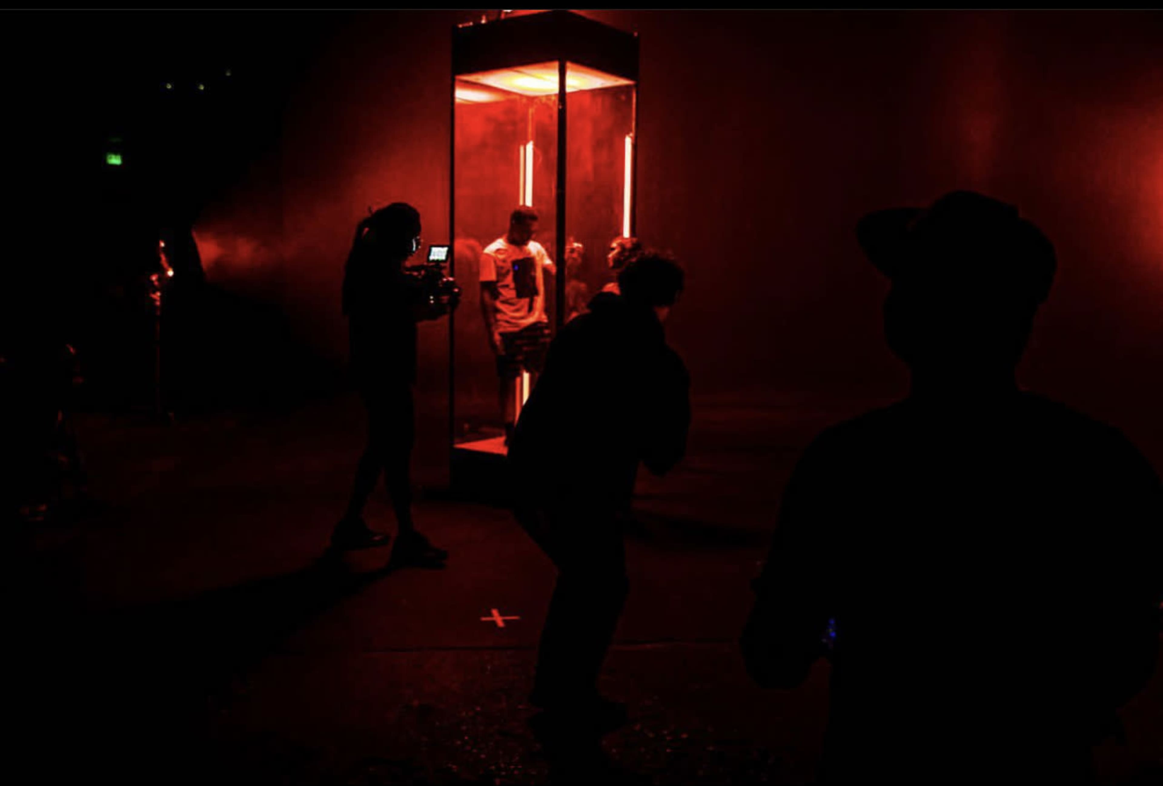 Two individuals are positioned in a red-lit studio setting, one inside a glass structure and another taking photos from outside.