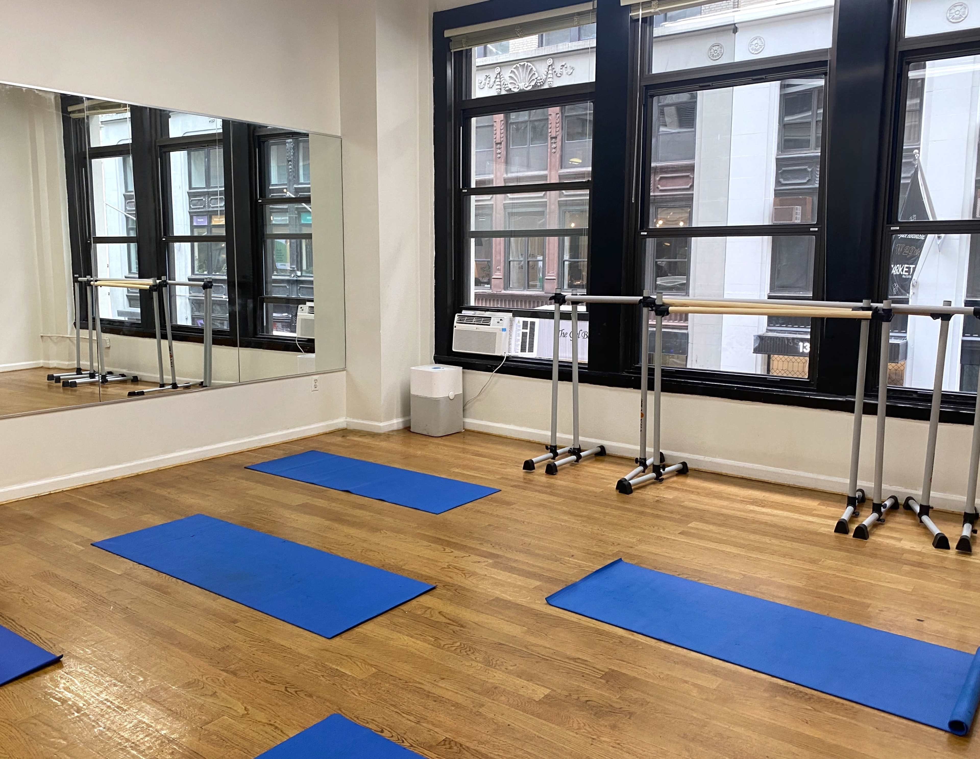 A brightly lit fitness studio with blue exercise mats on a wooden floor, mirrors on one wall, and ballet barres next to large windows.