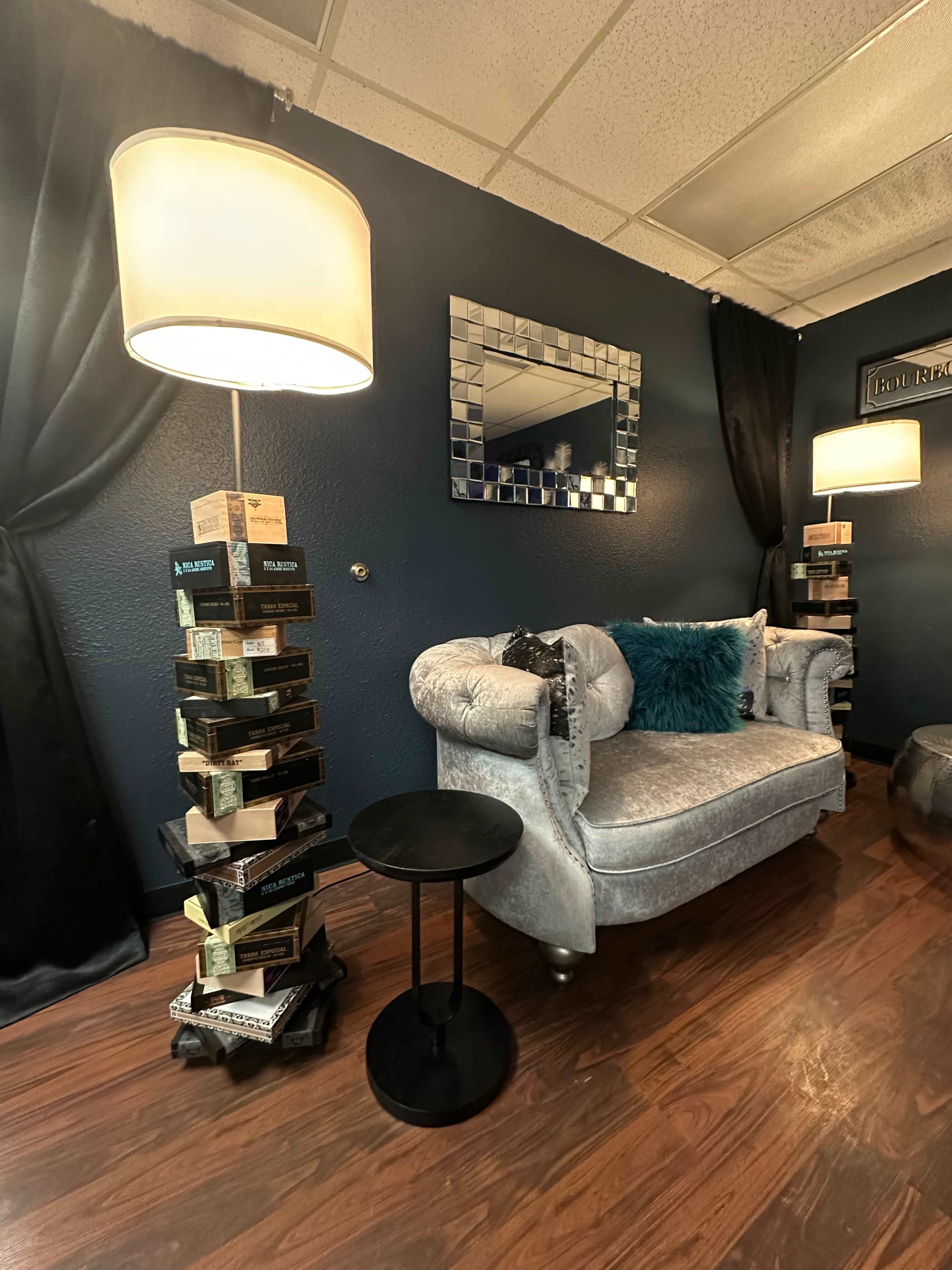 The image shows a cozy interior with a gray upholstered couch, a mirrored wall decoration, two standing lamps, and stacks of boxes on either side.