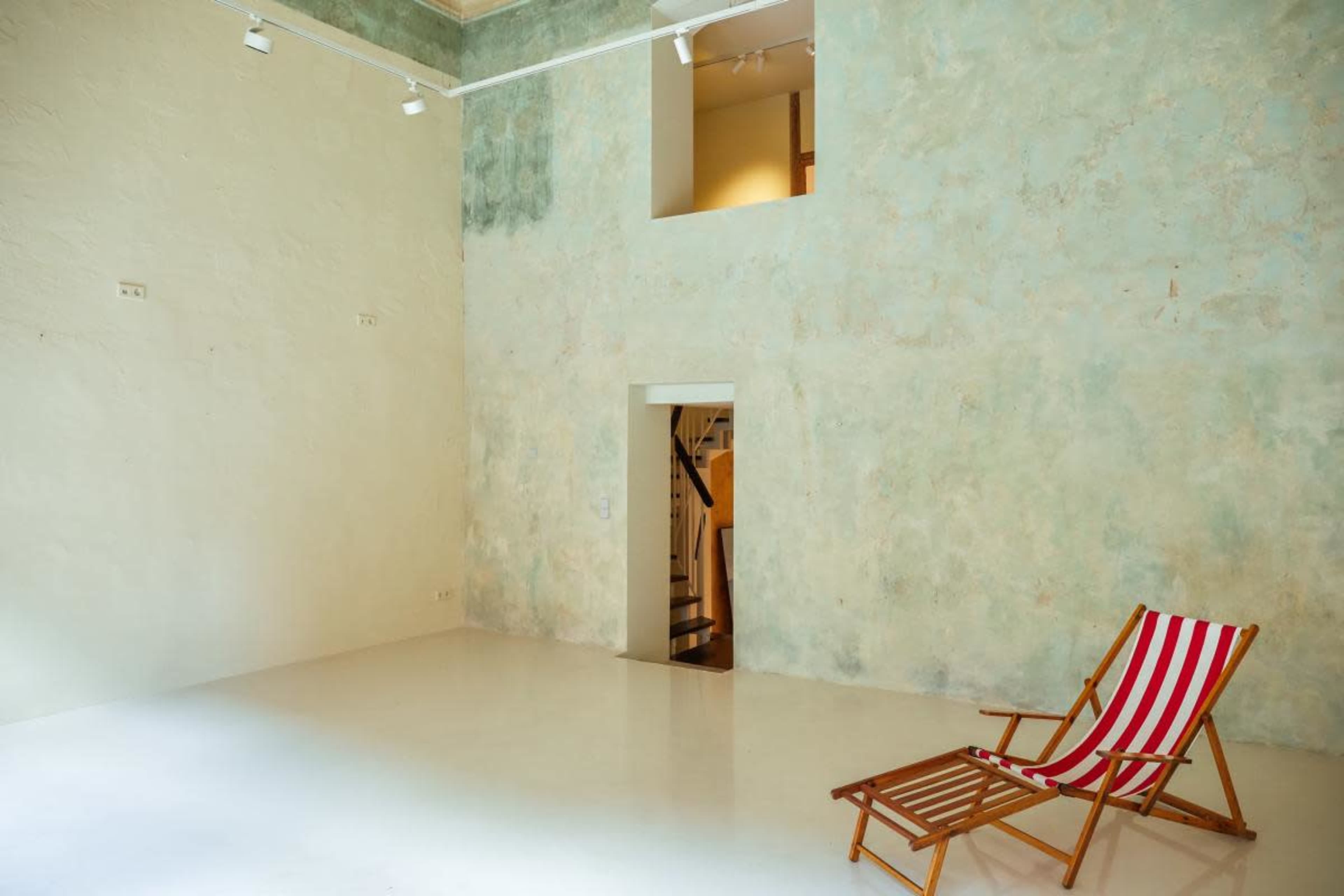 The image shows a spacious room with a textured, light green wall and a wooden deck chair with red and white stripes placed on a white floor.