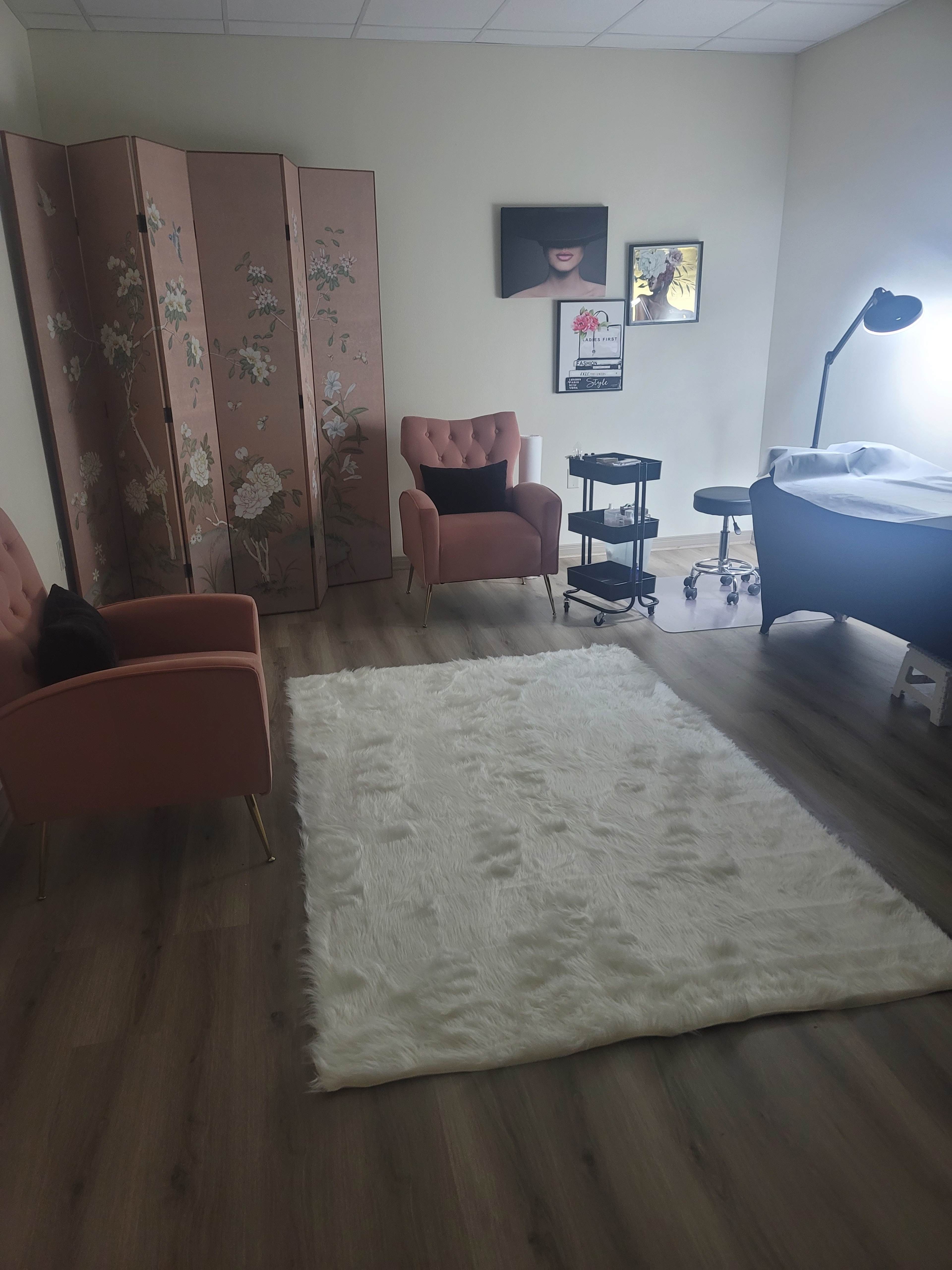 A softly lit room featuring two pink chairs, a decorative folding screen, a treatment table with a stool, and a large white rug.