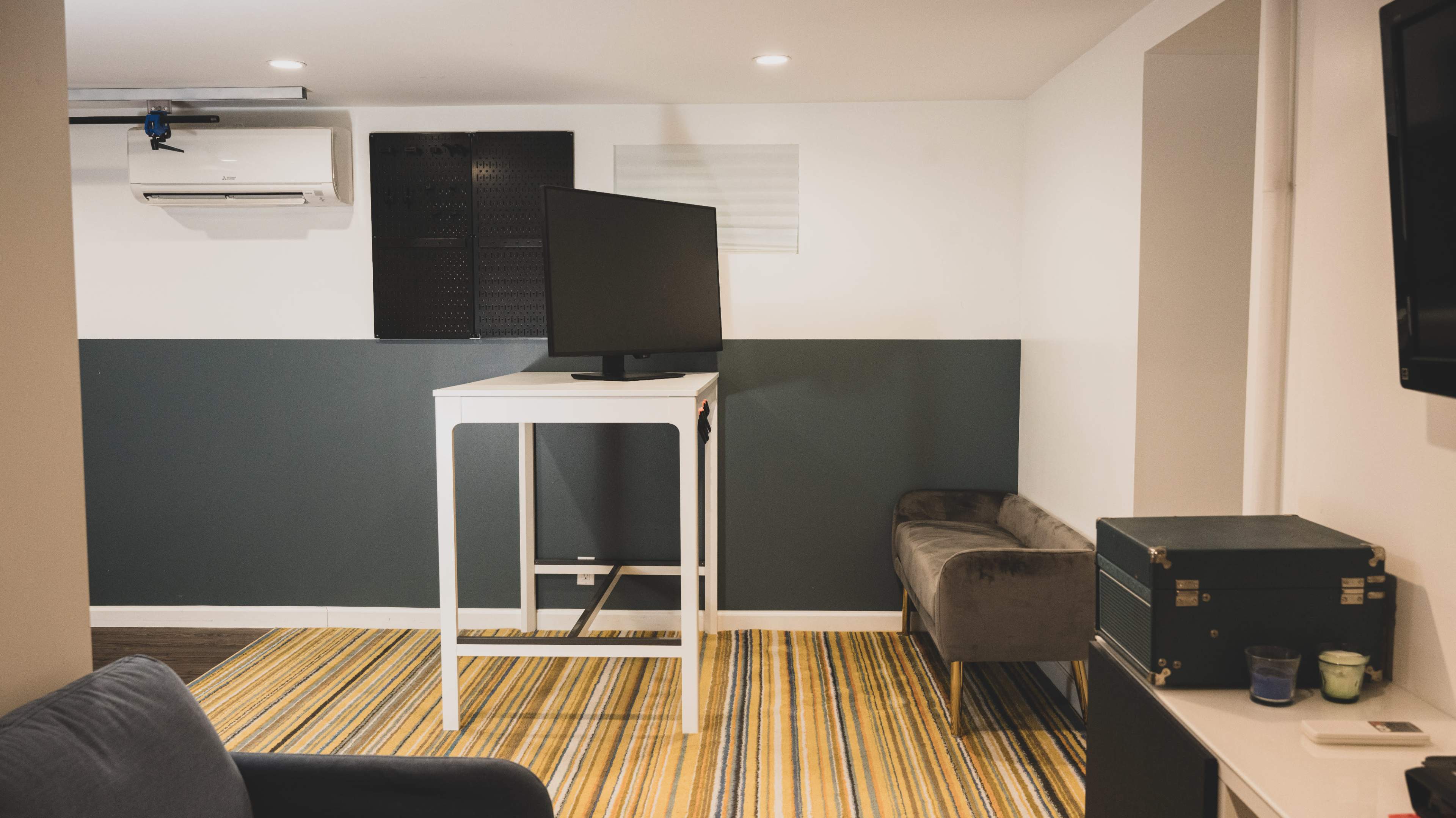 A small room features a standing white table with a television on top, a gray couch, and a striped rug on the floor.