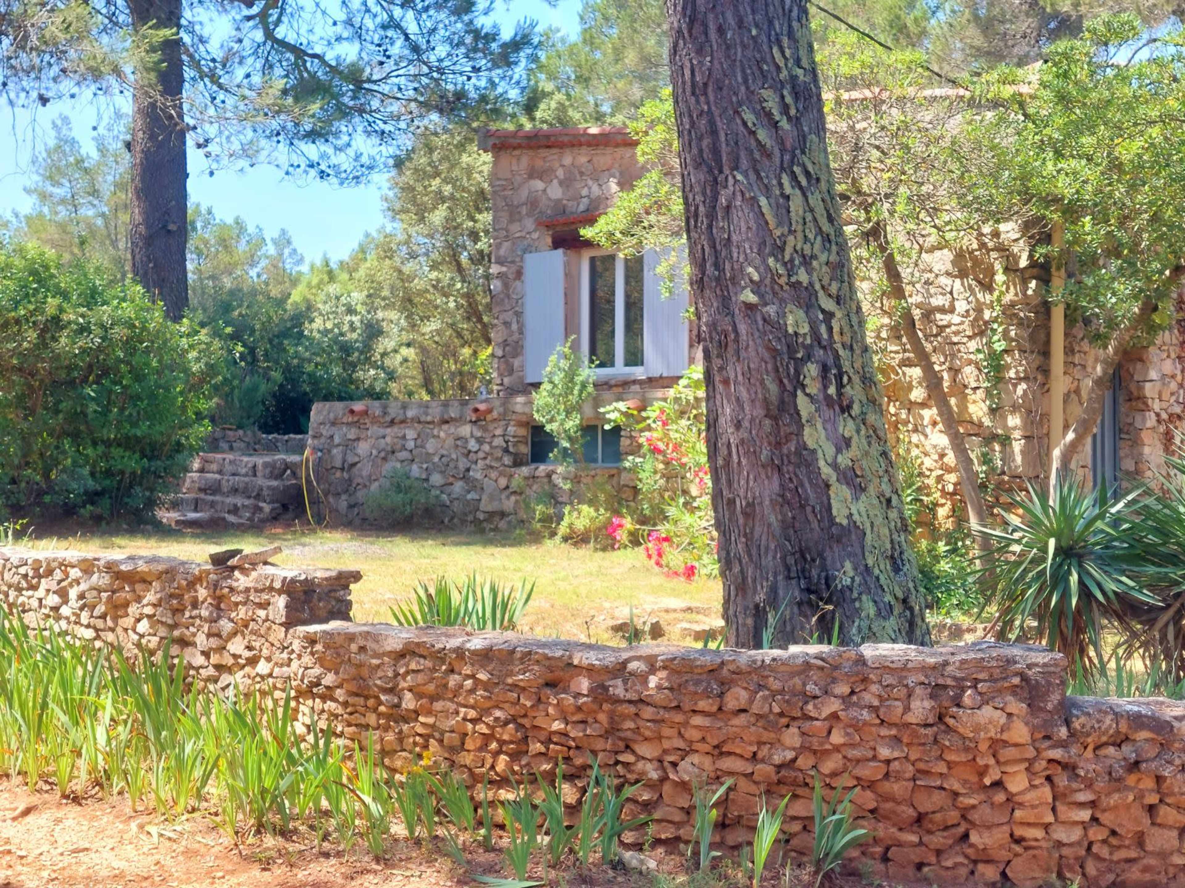 The image shows a rustic stone building surrounded by greenery and a low stone wall.